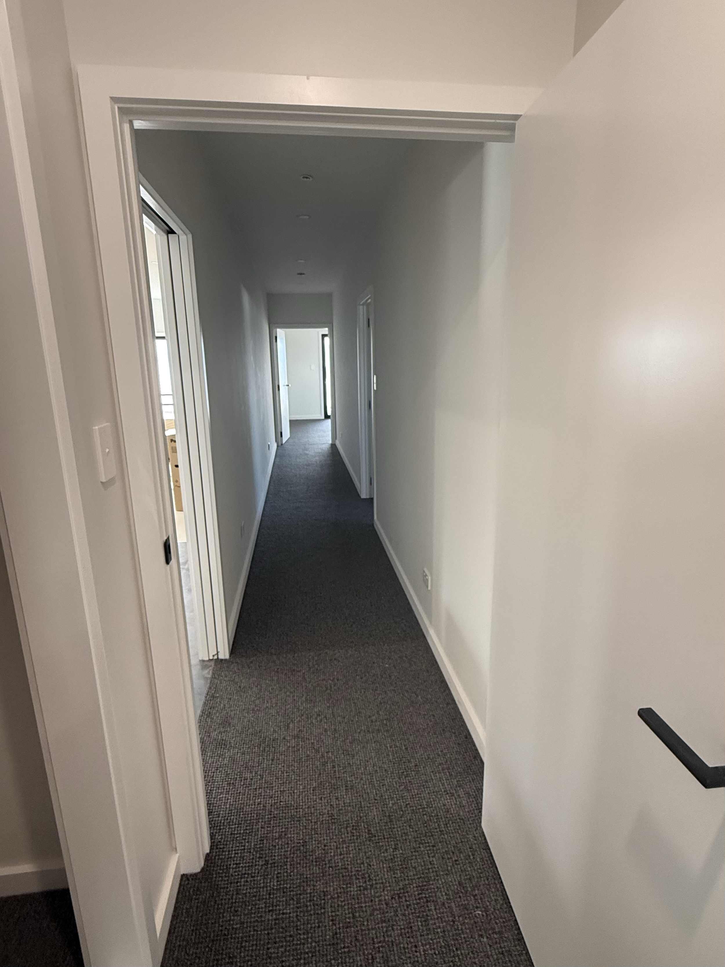 Empty hallway in a modern building with white walls, dark carpet, multiple doorways, and a staircase at the end.