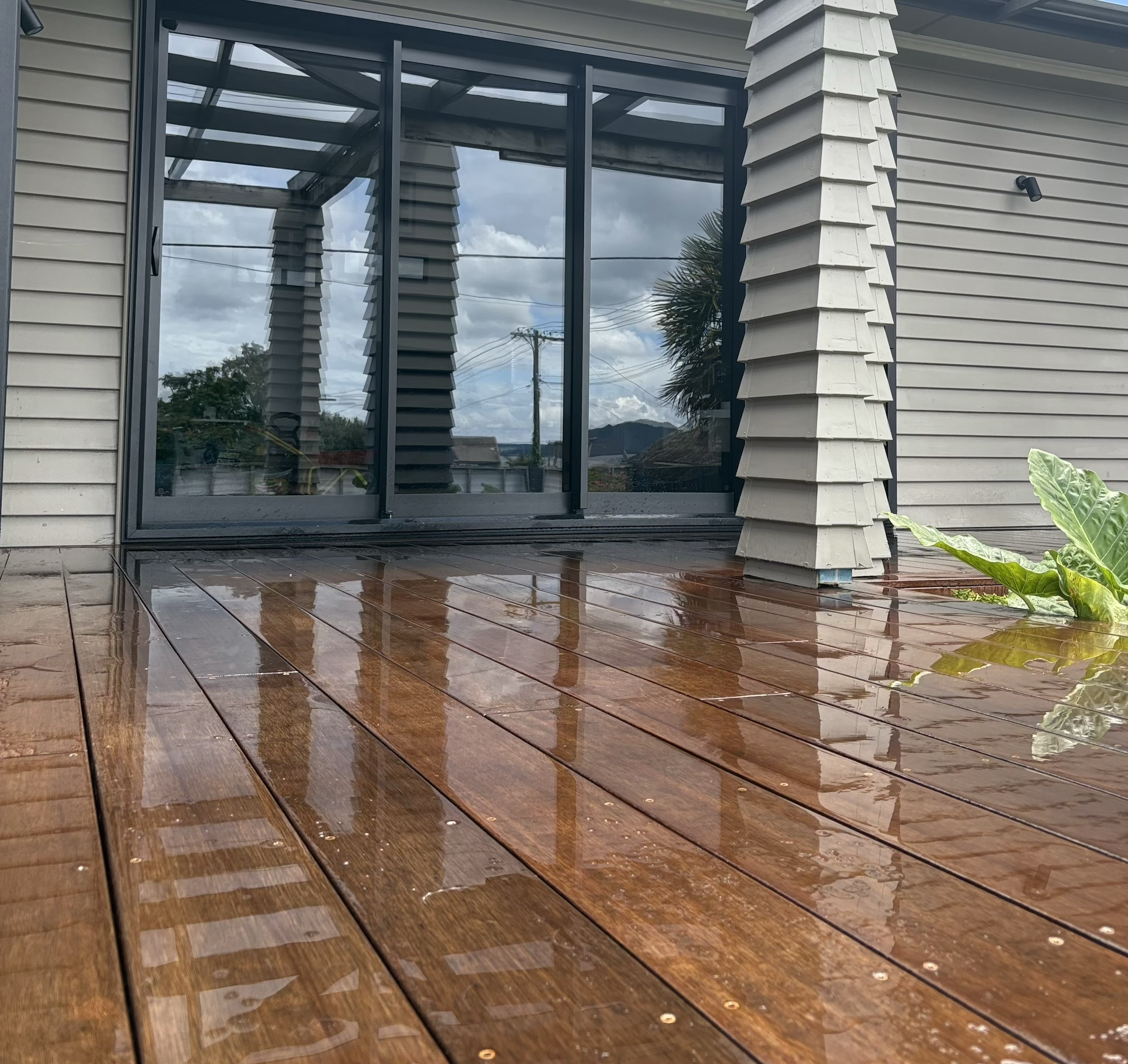 Wooden deck outside a house with sliding glass doors, gray siding, and a small green plant nearby under a cloudy sky.