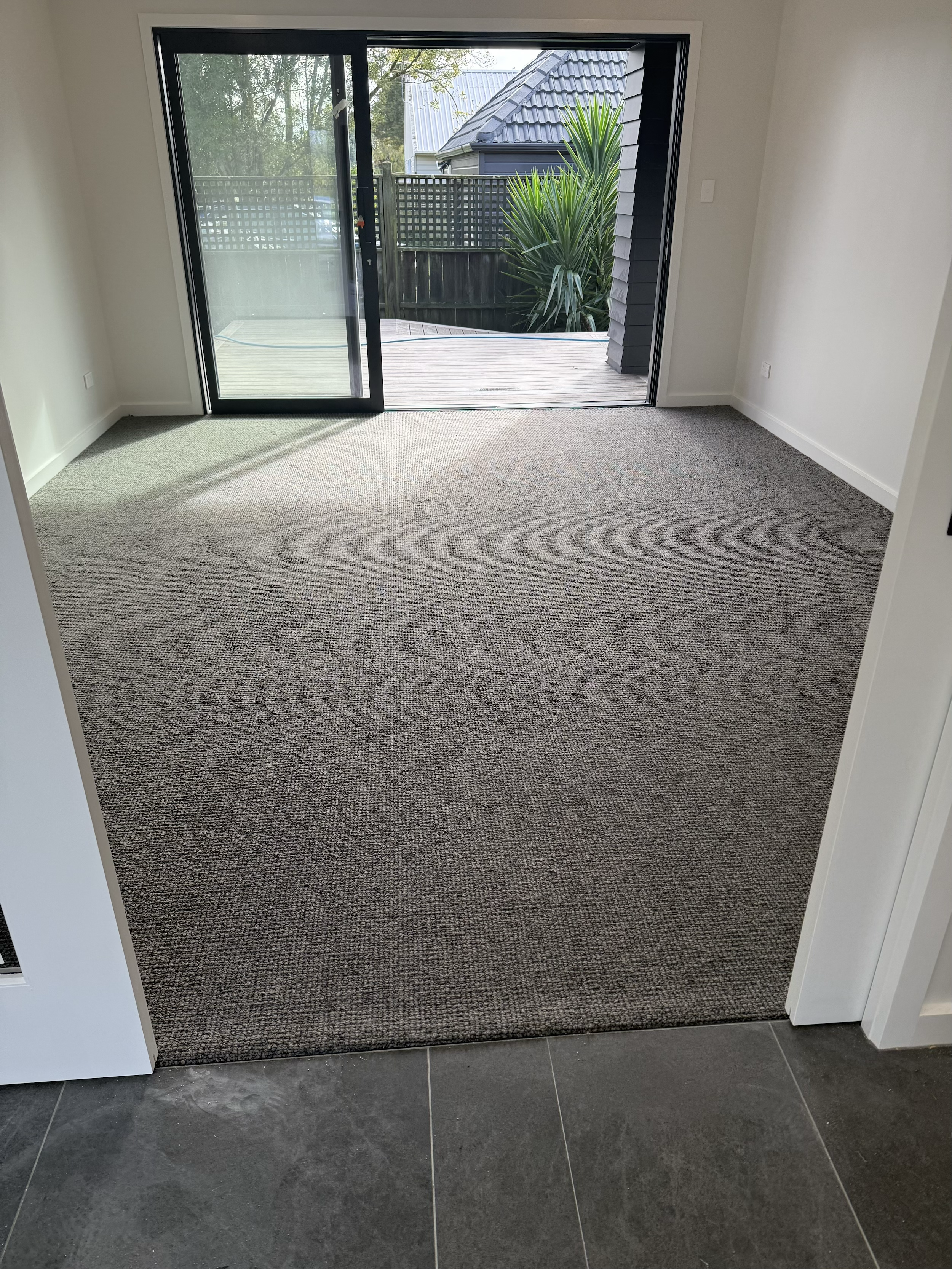 Interior view of a room with a sliding glass door leading to a small outdoor deck and a yard with green plants and trees, beige carpet flooring, and white walls.