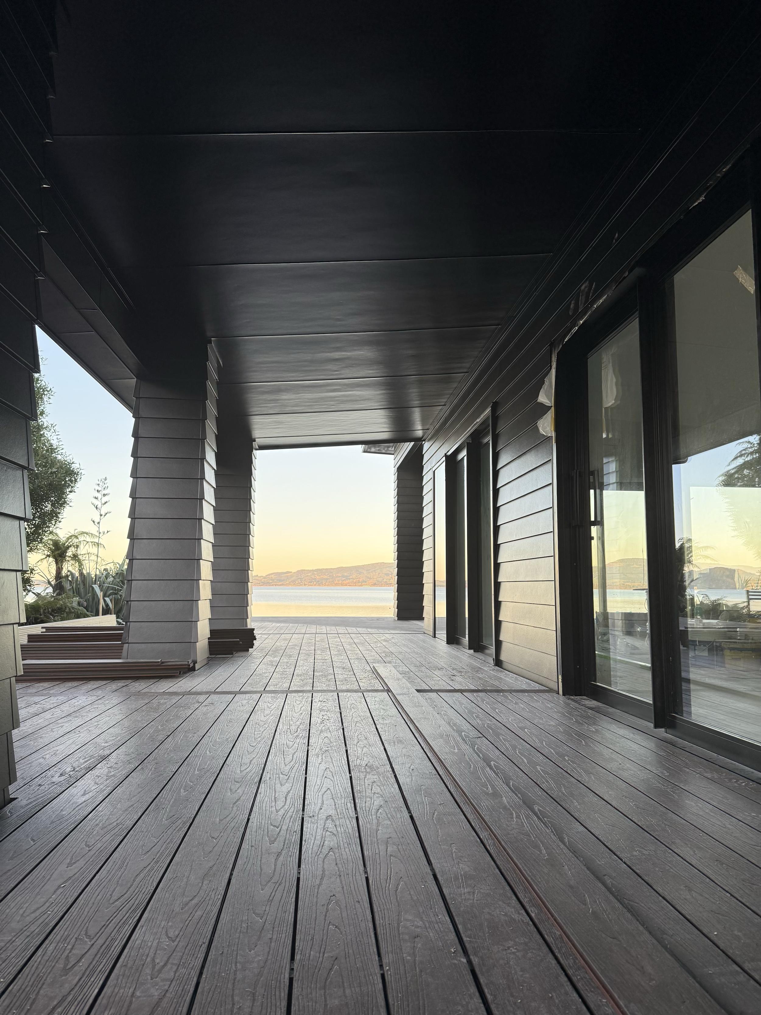 View from a spacious deck with wooden flooring overlooking a body of water and distant land, with a modern house's exterior wall with glass sliding doors on the right and structural columns on the left.