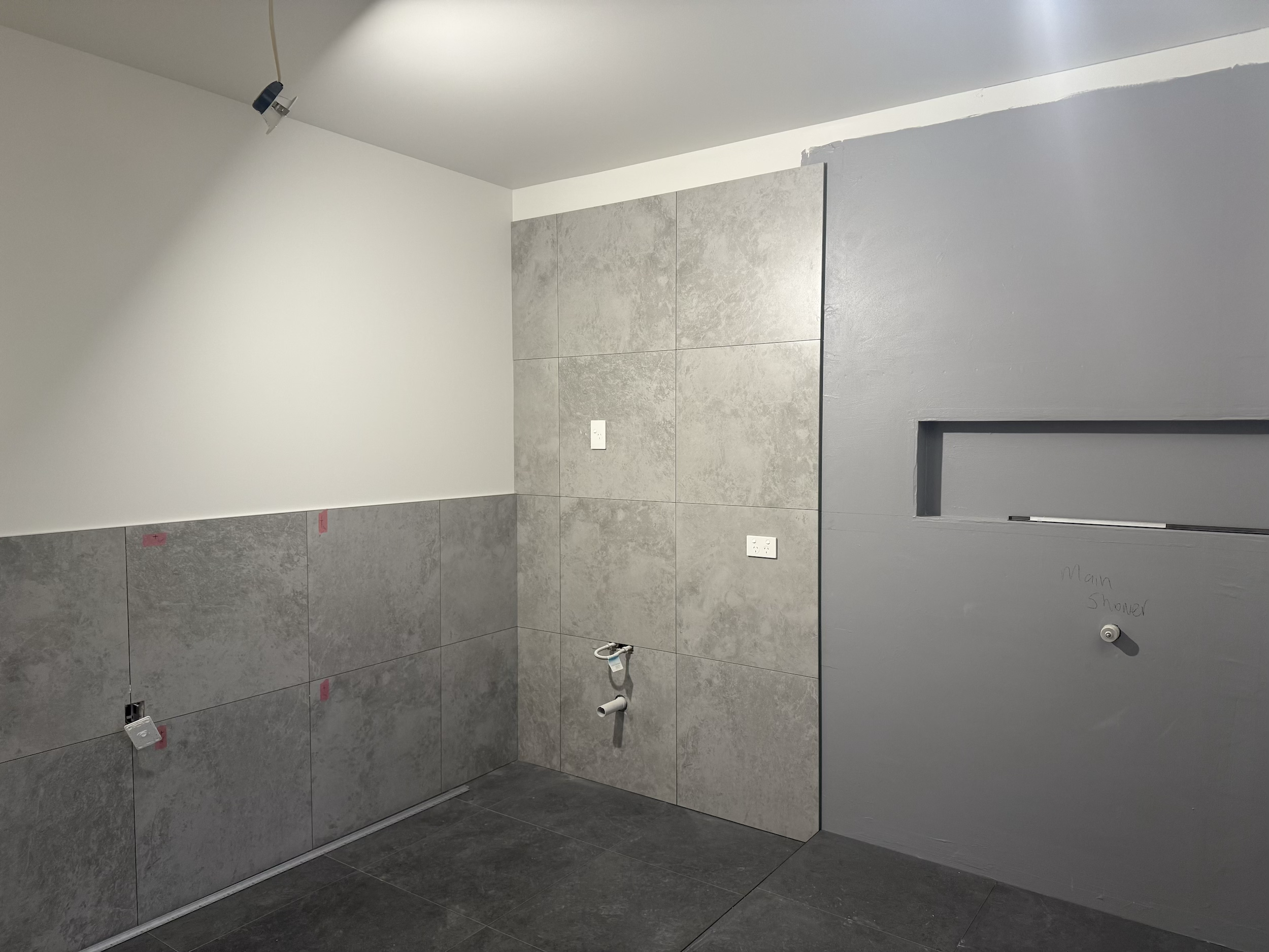 Bathroom under construction with gray and white tiles, electrical outlets, and plumbing fixtures.