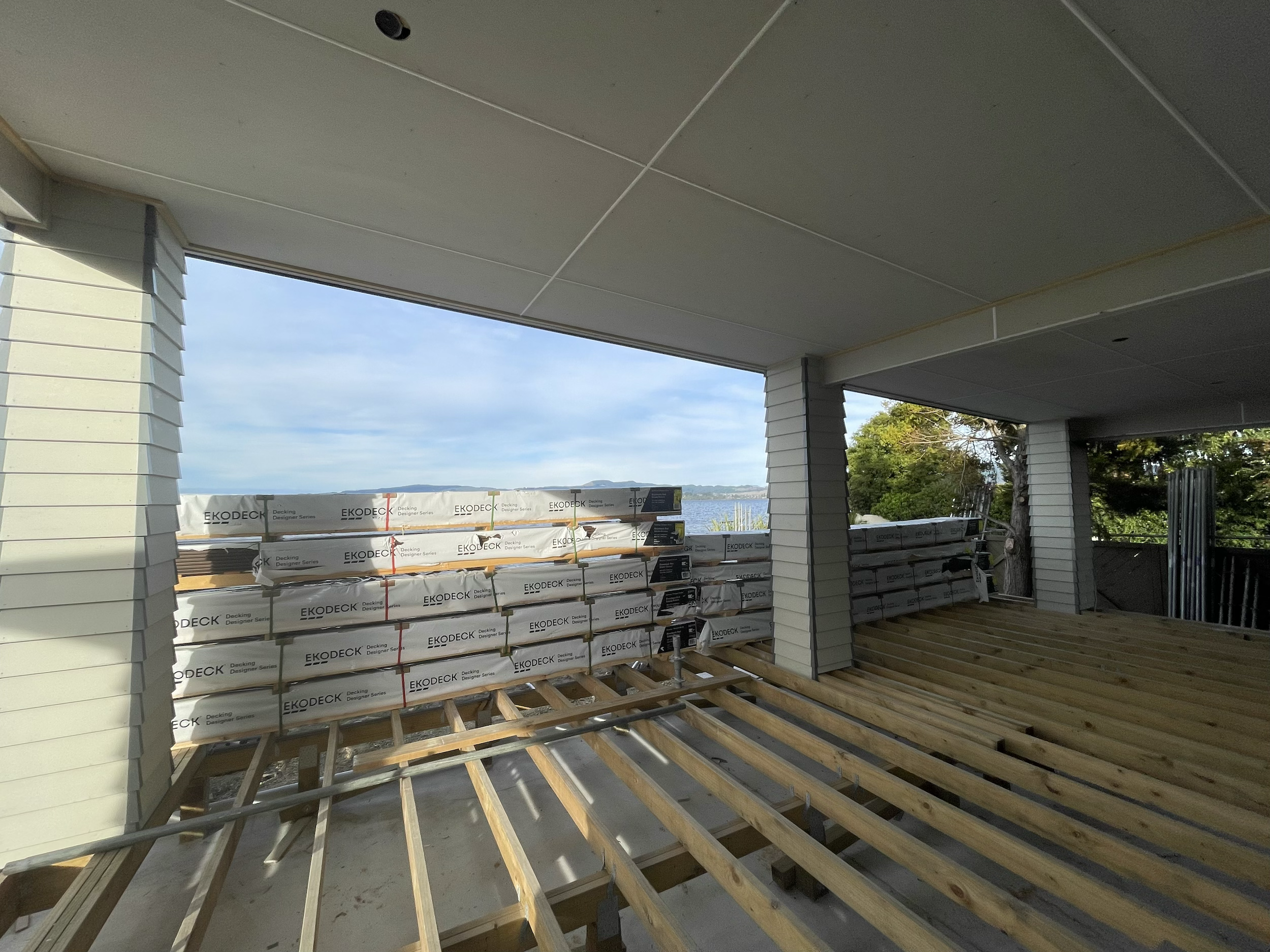 Construction site with a partially built wooden deck overlooking a body of water, with stacks of roofing materials and green trees in the background.