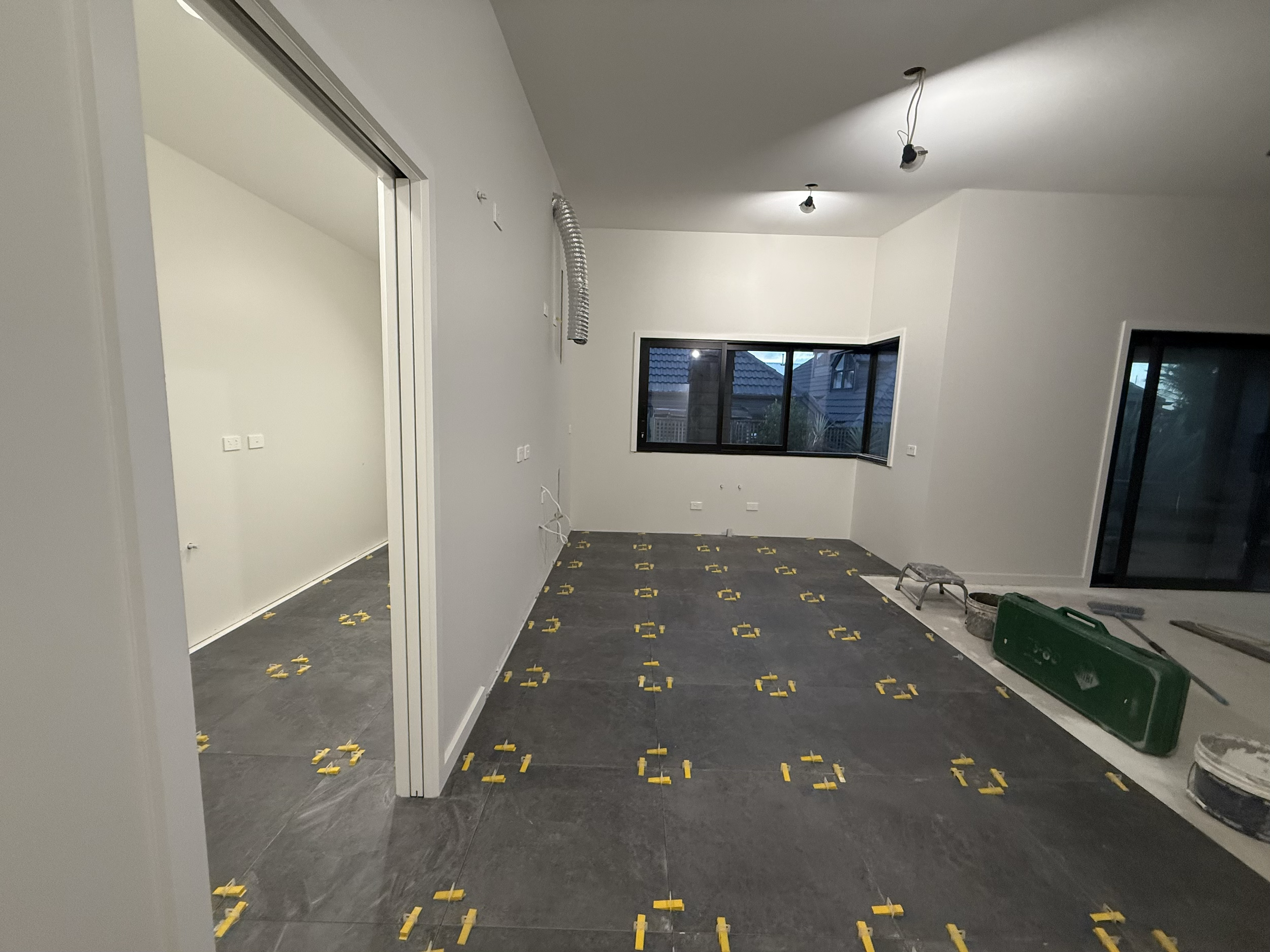 Interior of a room under renovation with dark floor tiles and yellow spacers, white walls, and a large window showing an outdoor scene. 