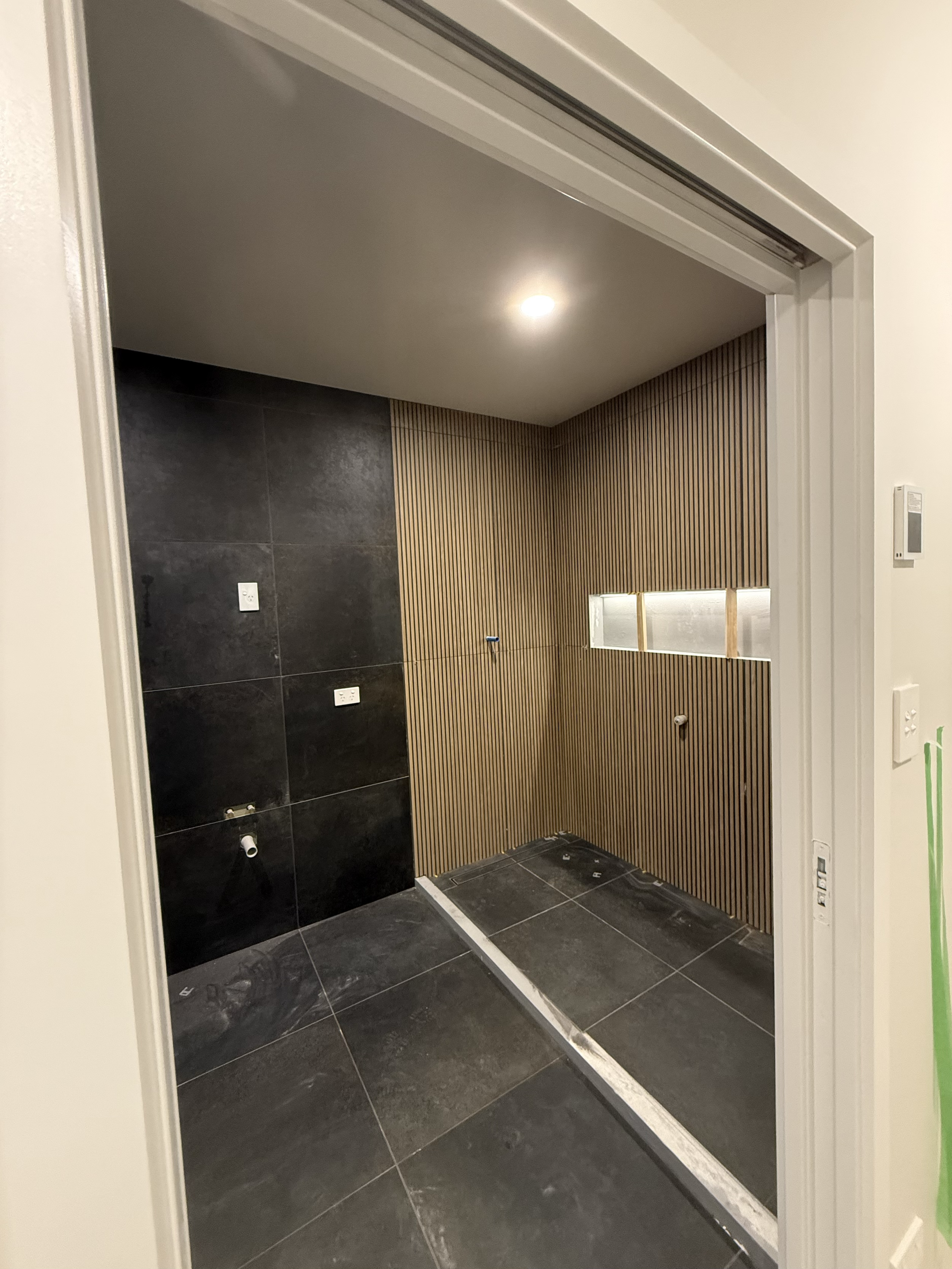 An unfinished bathroom with black tiled floor and black tiled wall on the left. The right wall is made of vertical wooden slats with a horizontal window. There are electrical outlets and plumbing fixtures installed but no fixtures yet. The ceiling ha