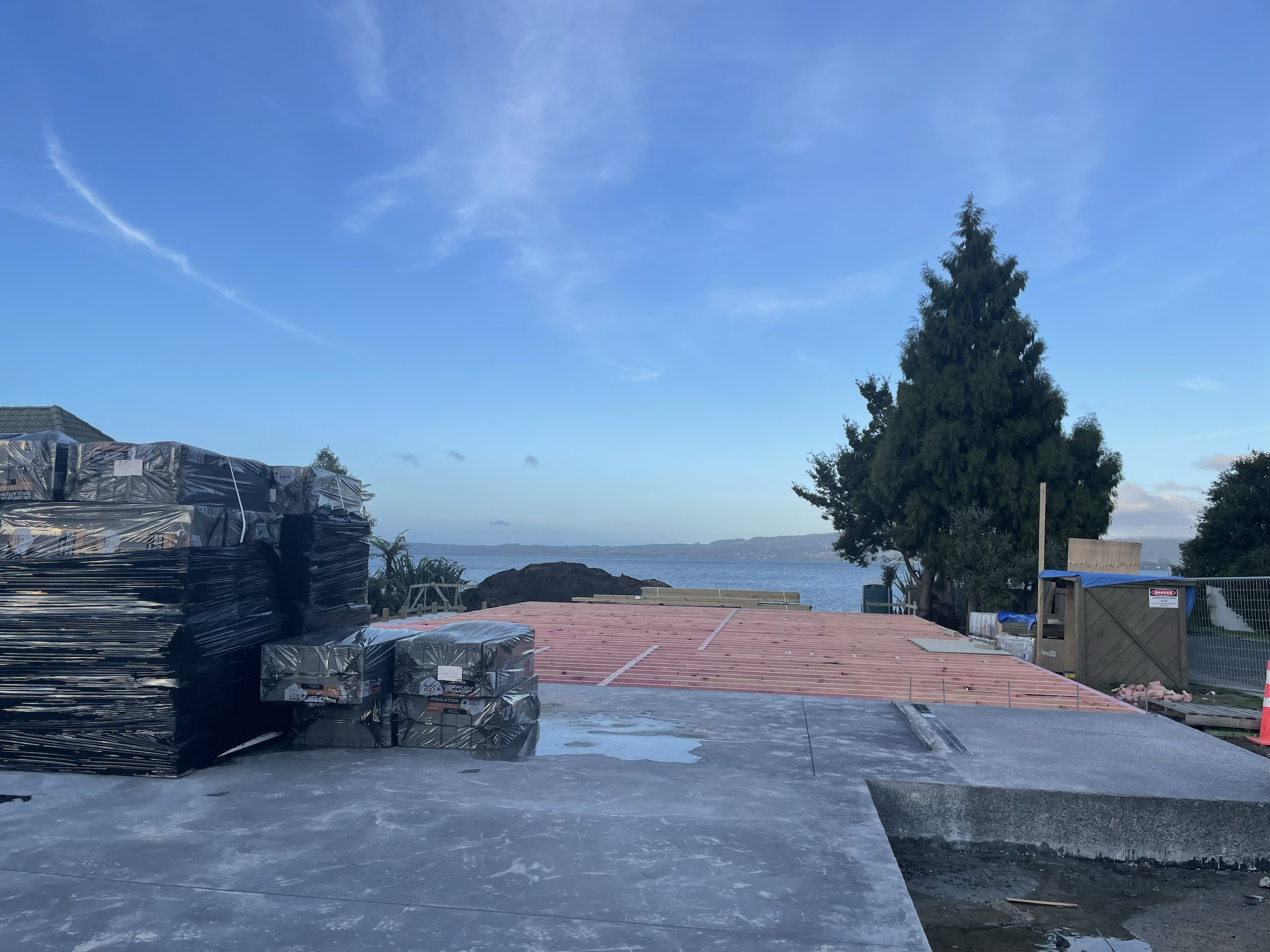 Construction site overlooking a body of water, with stacks of building materials and a partially built structure on a sunny day.