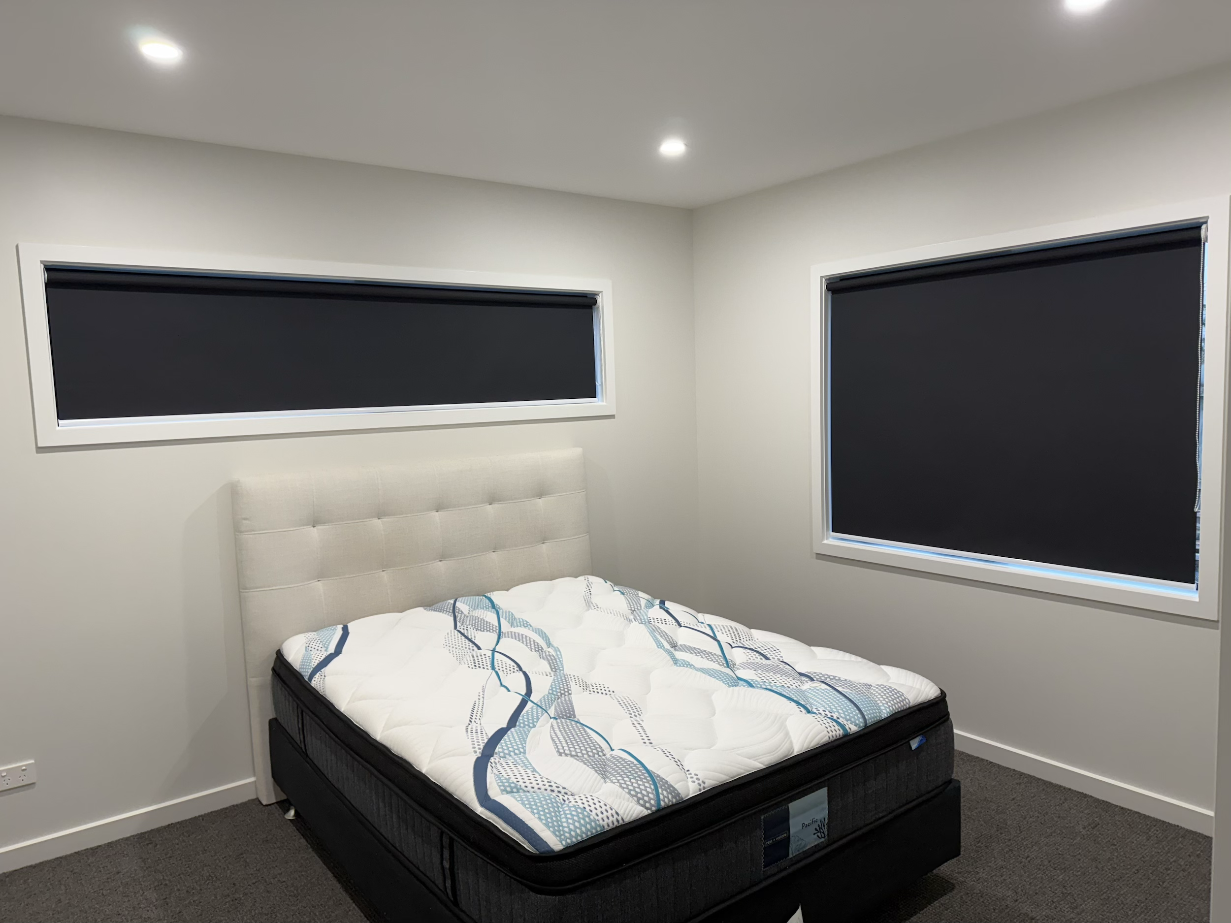 A bedroom with a mattress on a black bed frame, beige headboard, white walls, two black window shades, and ceiling lights.
