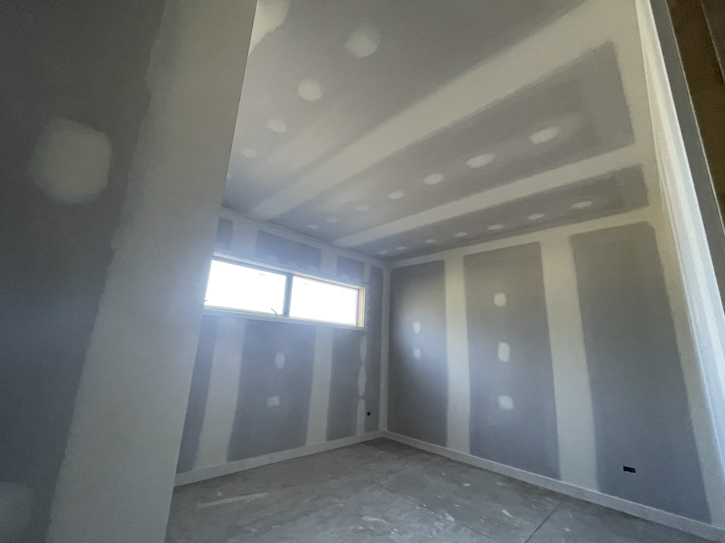 An unfinished room with drywall installed, seams taped, and joint compound ready for painting, with a horizontal window letting in natural light.
