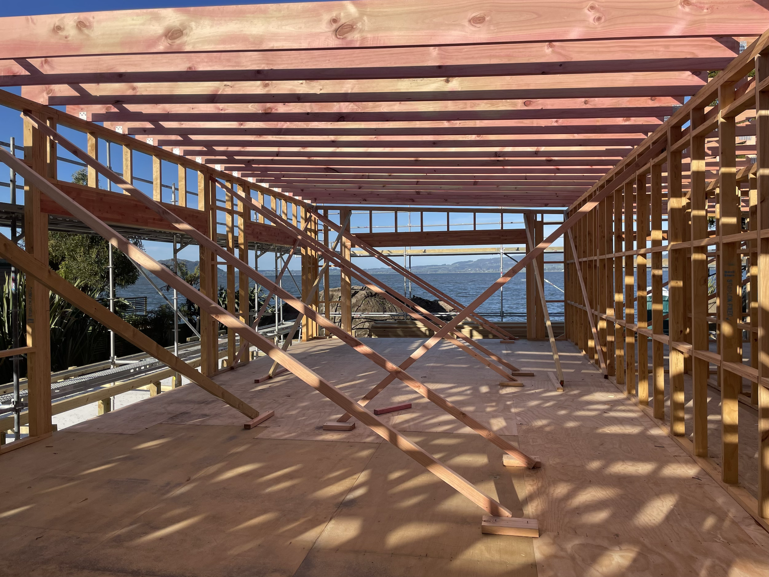 The image shows a partially built wooden house frame with exposed beams and walls, overlooking a body of water with a rocky shoreline and distant mountains. The construction is outdoors with bright sunlight and clear skies.