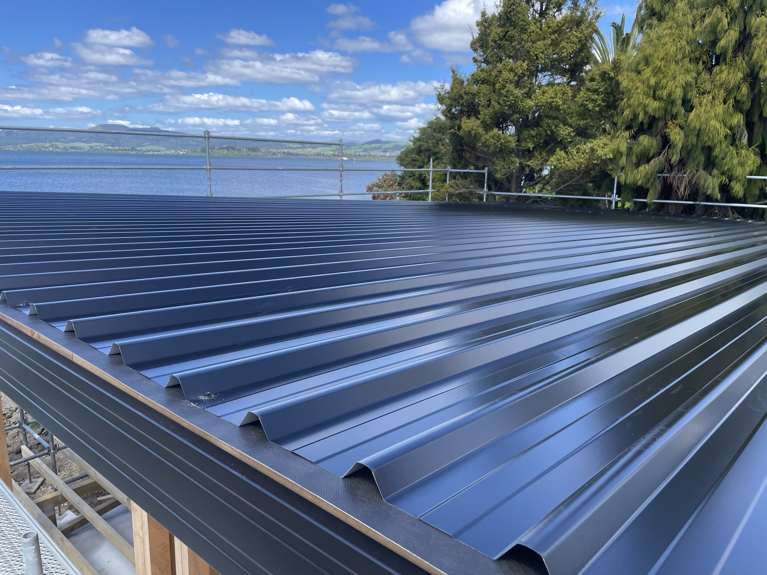 A new dark gray or black metal roof on a building under construction, with a lake, trees, and blue sky with clouds in the background.