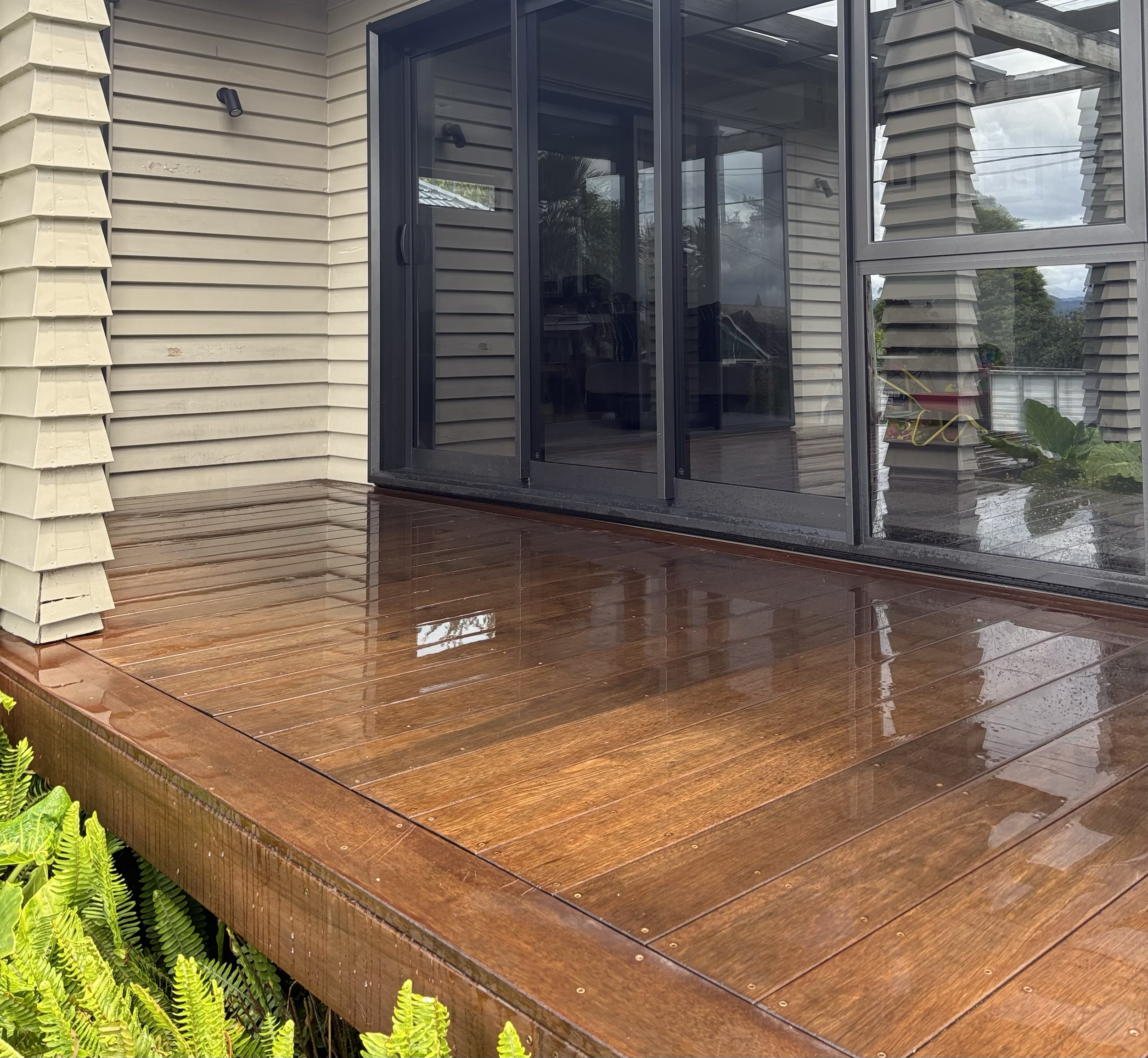Wooden deck outside a house with glass sliding doors, showing reflections of the house and surrounding landscape.