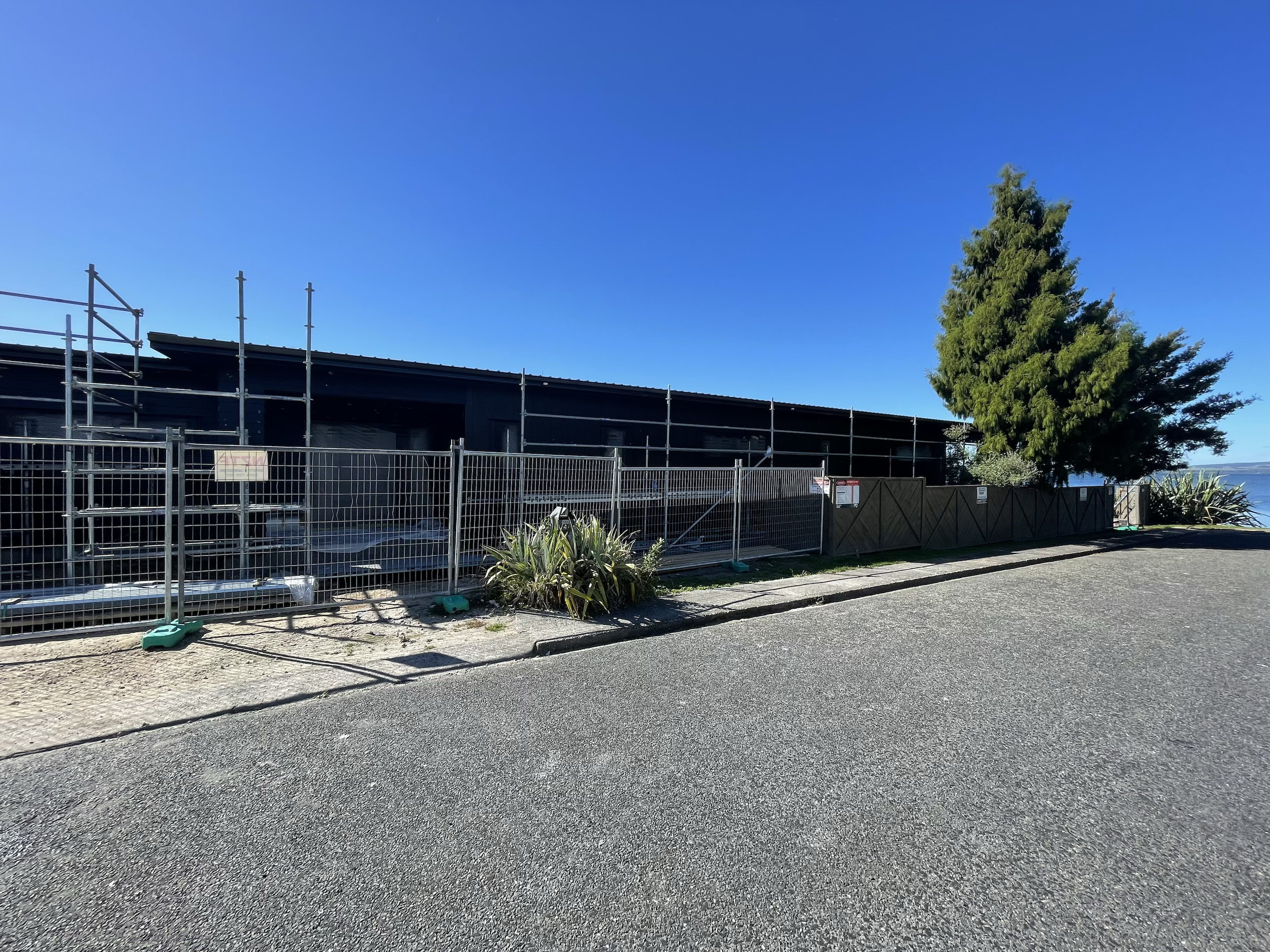 A construction site next to a sidewalk with fencing, a building under renovation, a tree, and a glimpse of water in the background under a clear blue sky.