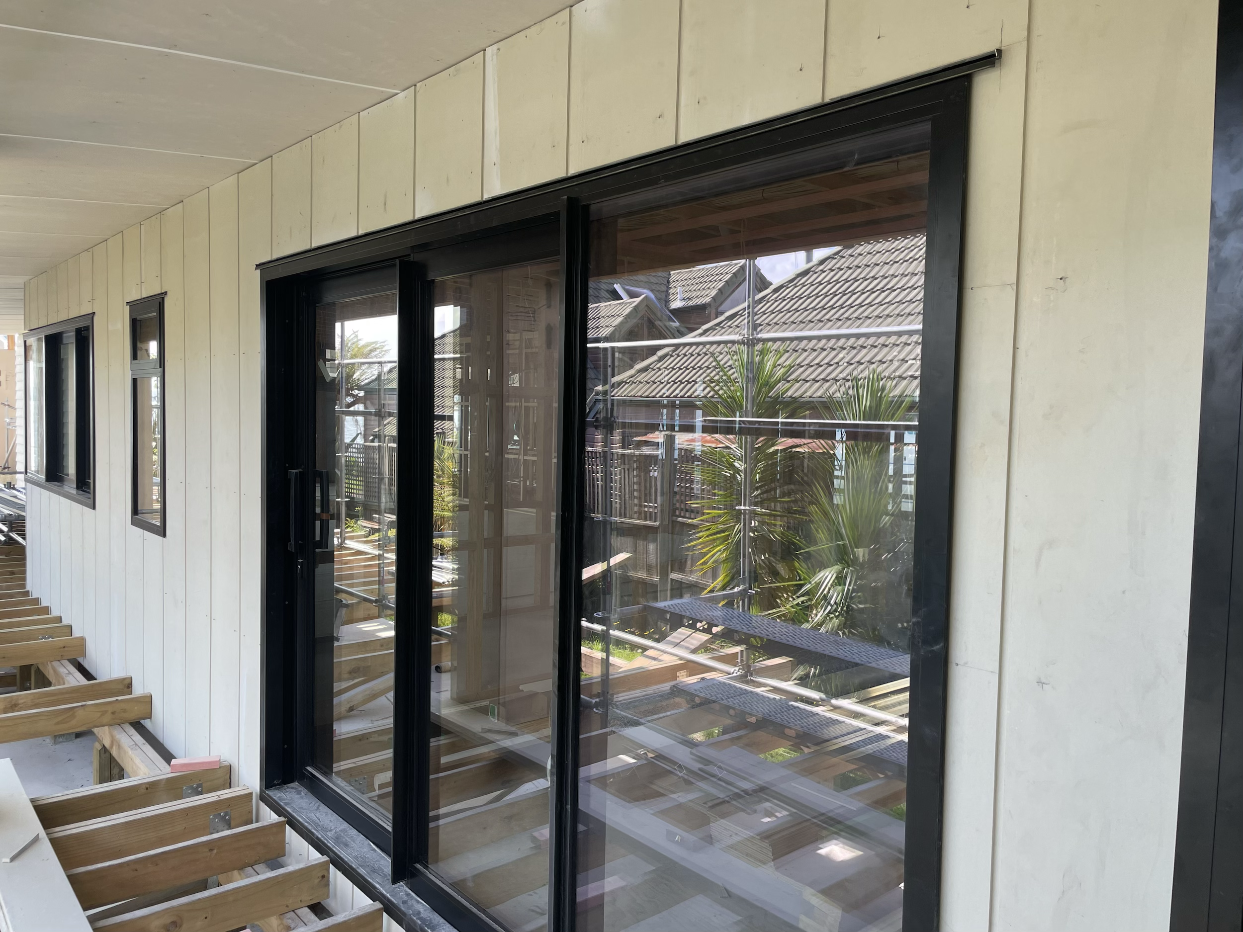 Exterior view of a building under construction with a sliding glass door and multiple windows, scaffolding, palm trees, and rooftops visible outside.