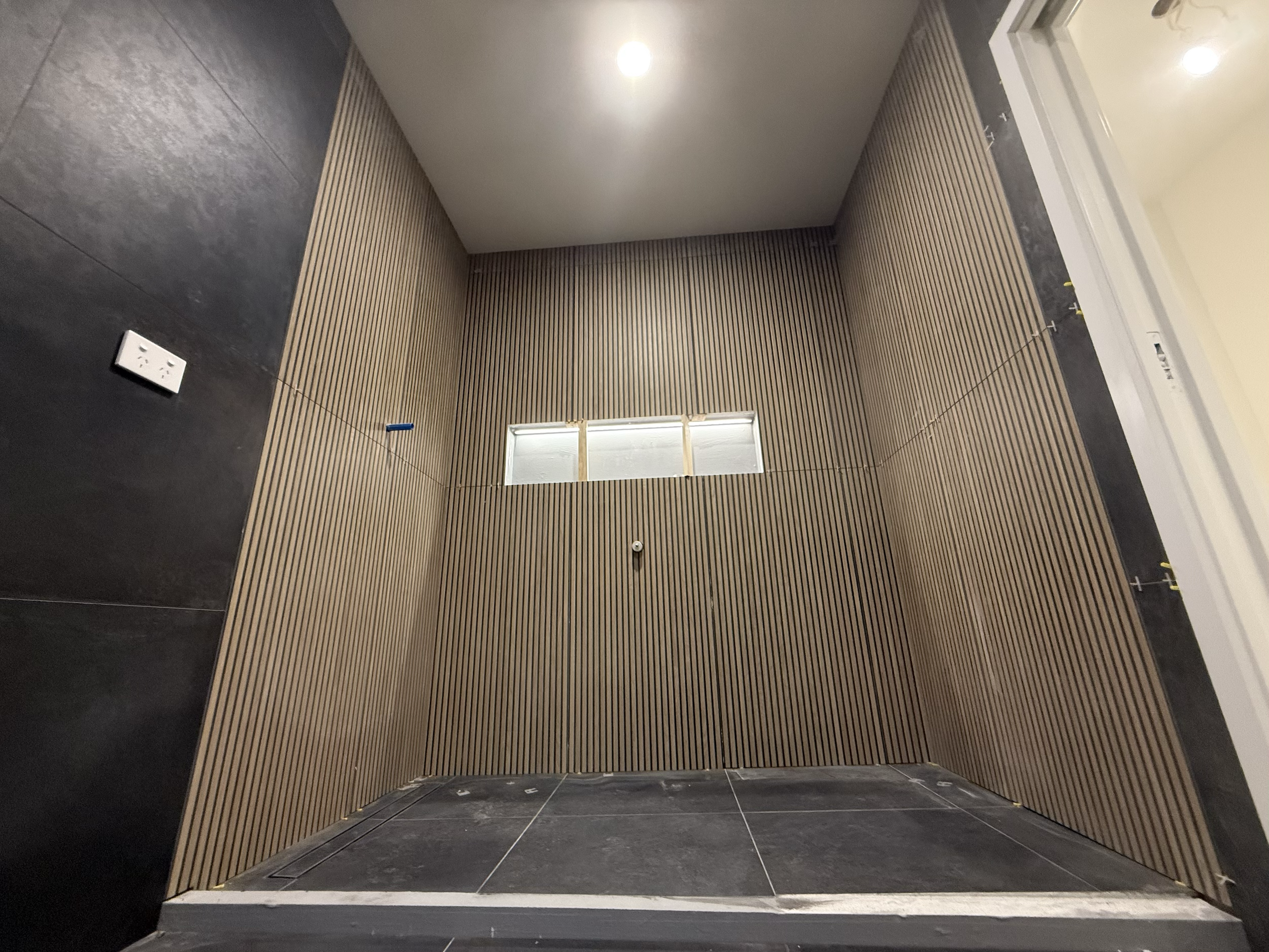 A room under construction with wooden slat walls, dark tiled flooring, and a small horizontal window with blinds on the back wall. An electrical outlet is on the left wall, and some wiring or string is suspended across the room.