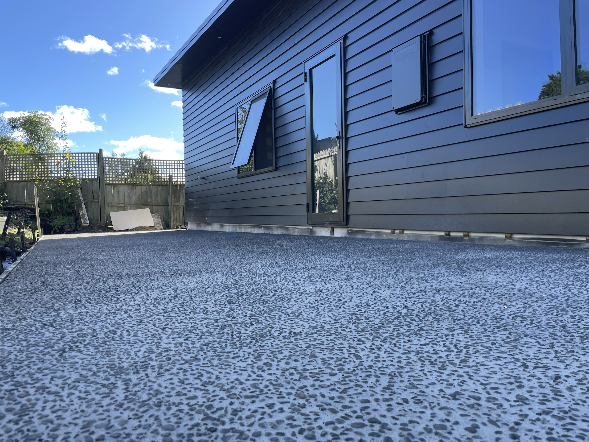 Newly installed textured concrete porch next to a dark gray house with black shingled siding, windows, and a door, with a fenced backyard and blue sky.
