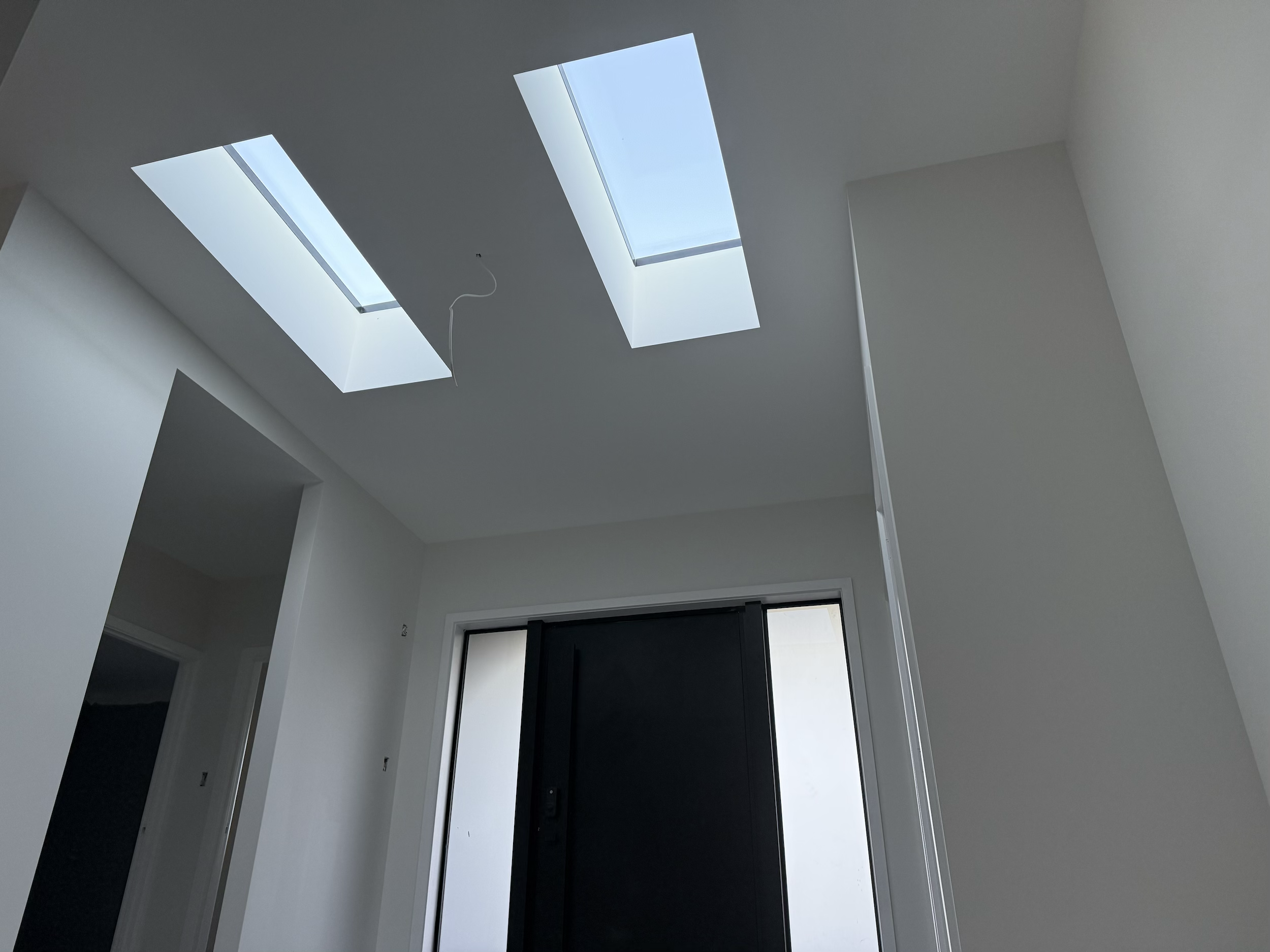 Interior view of a modern room with a white ceiling featuring two skylights, one with a hanging wire. The room has white walls, a black front door, and an open doorway.