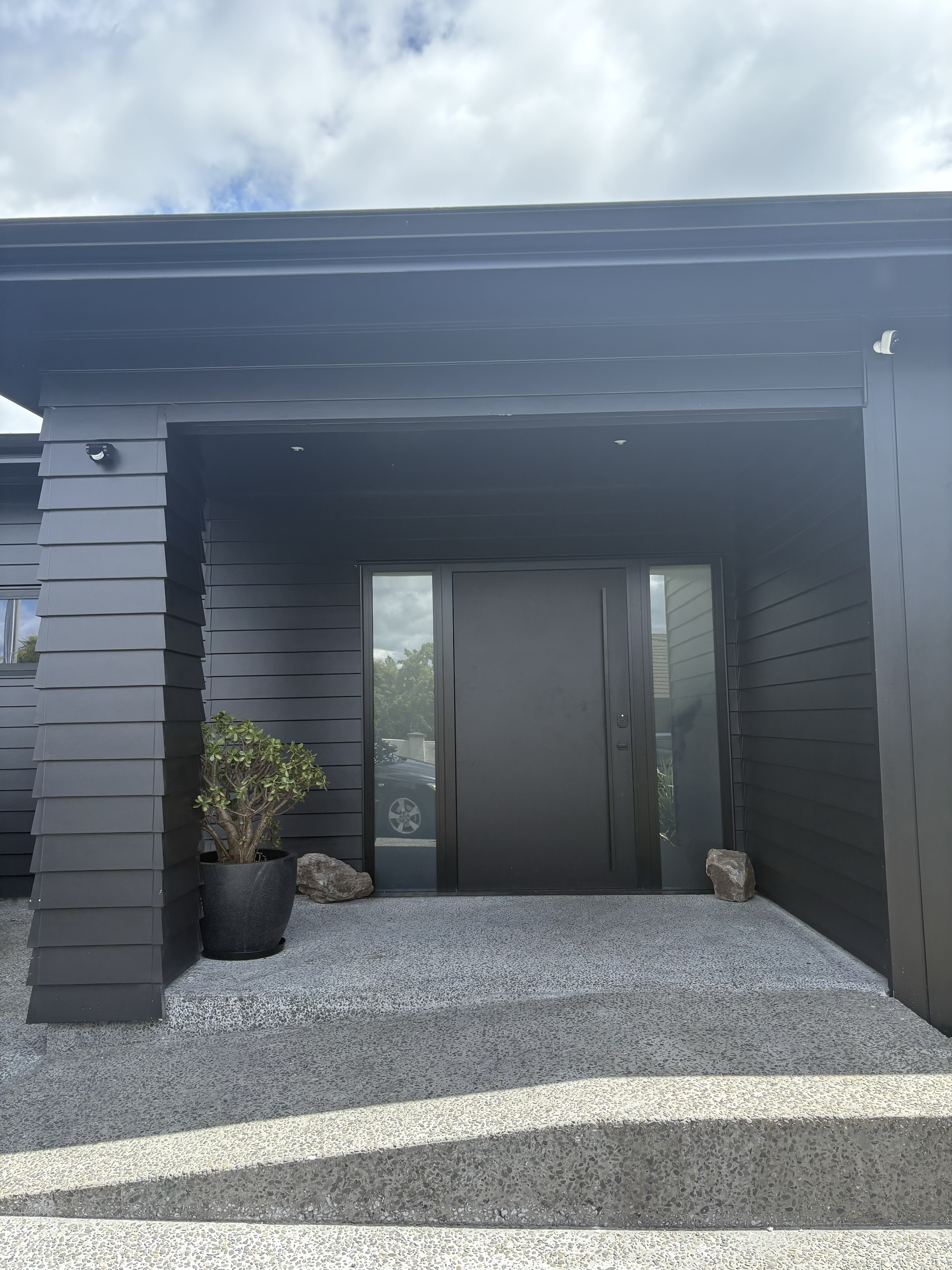 The front exterior of a modern black house entrance with a black door, glass side panels, potted plant, and stone accents on the staircase.