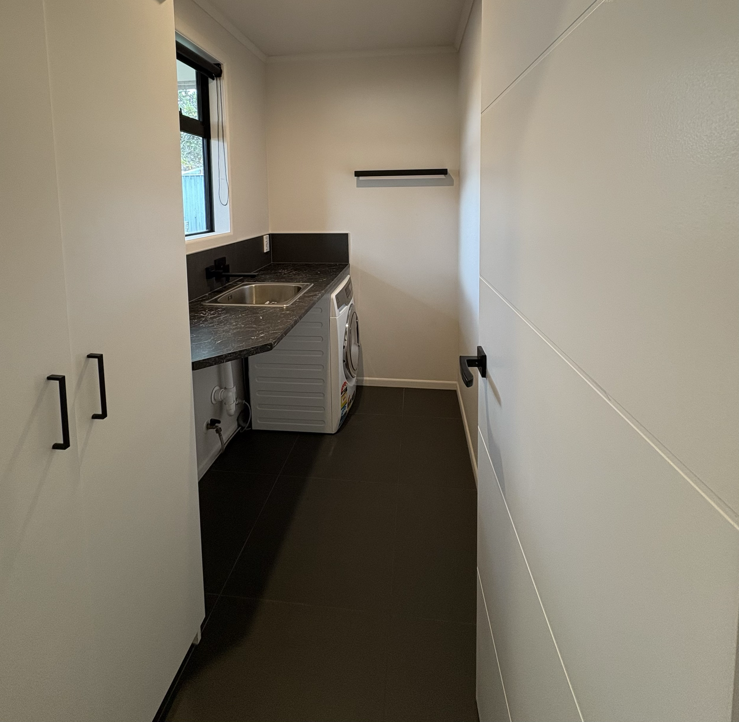 Small laundry room with a washing machine, a sink, a window, a black countertop, white cabinets, and a black shelf on the wall.