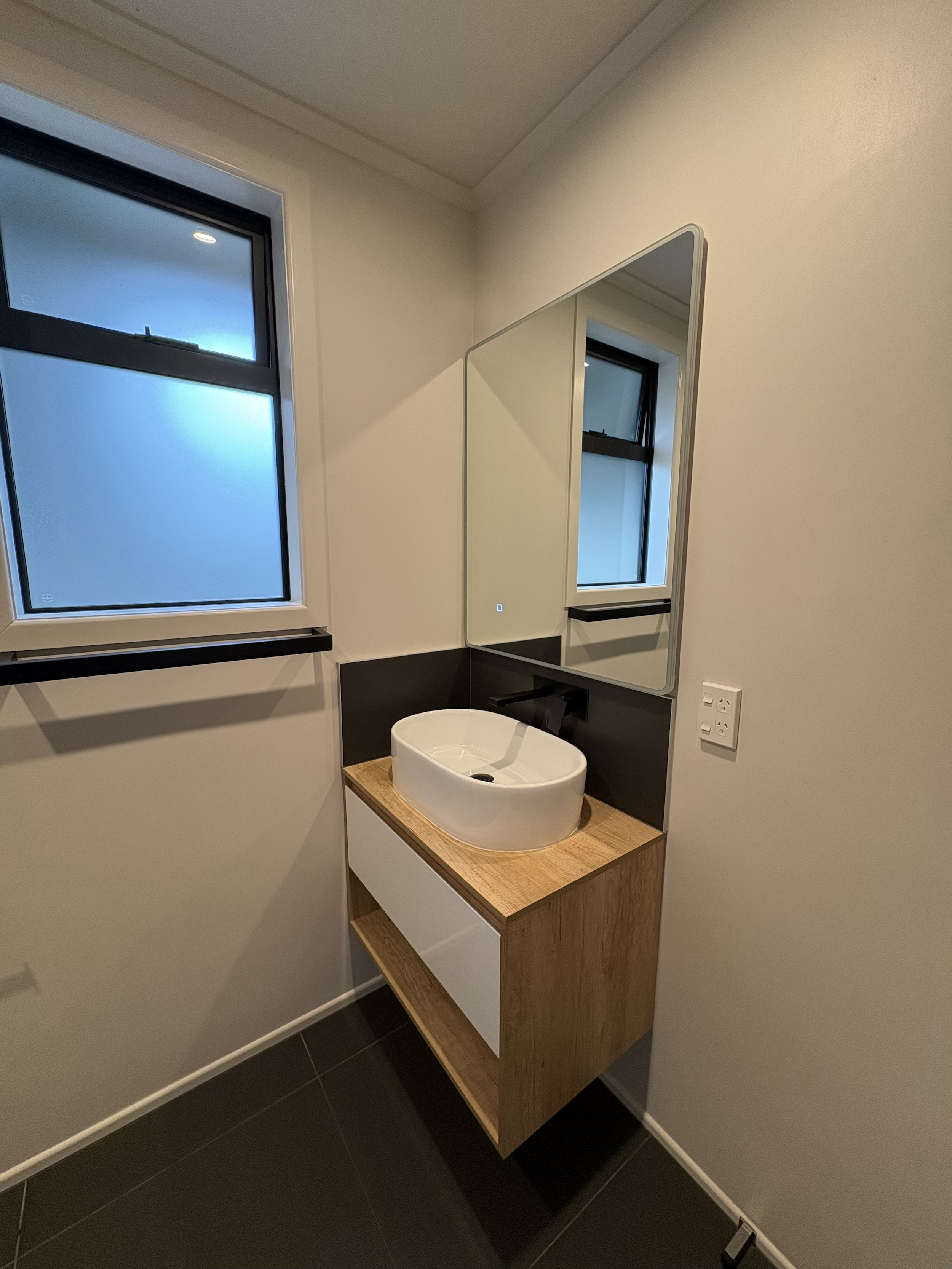 A modern bathroom with a round basin sink on a wooden vanity, a large wall mirror, and frosted windows, with black trim and a black tile backsplash.