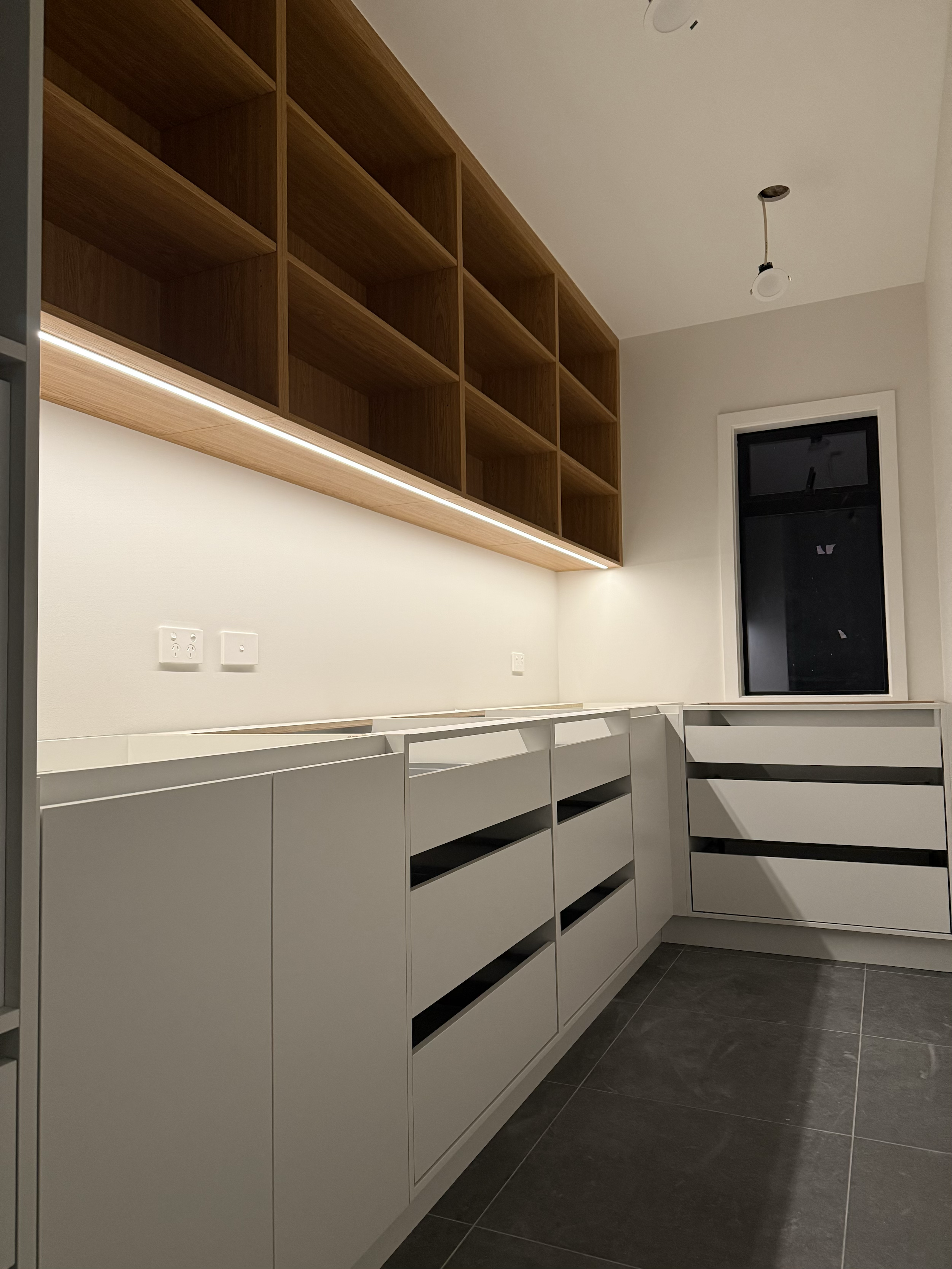 Modern kitchen or pantry with white cabinets, dark tiled floor, and open wooden shelves on the wall. A window is in the corner, and there are electrical outlets on the wall.