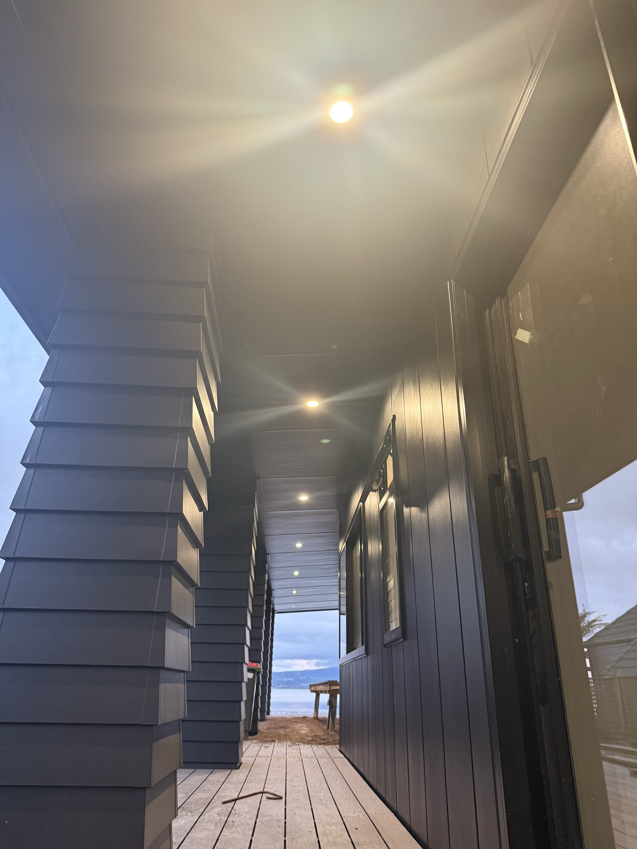 Photo of a covered porch with gray siding, wooden flooring, and bright ceiling lights, overlooking a body of water and mountains in the distance.