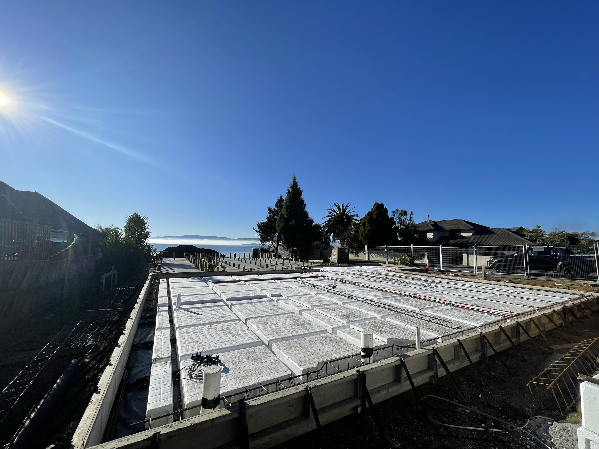 Construction site with concrete foundation and insulation panels, fenced area, nearby houses, palm trees, and a clear blue sky with the sun shining.