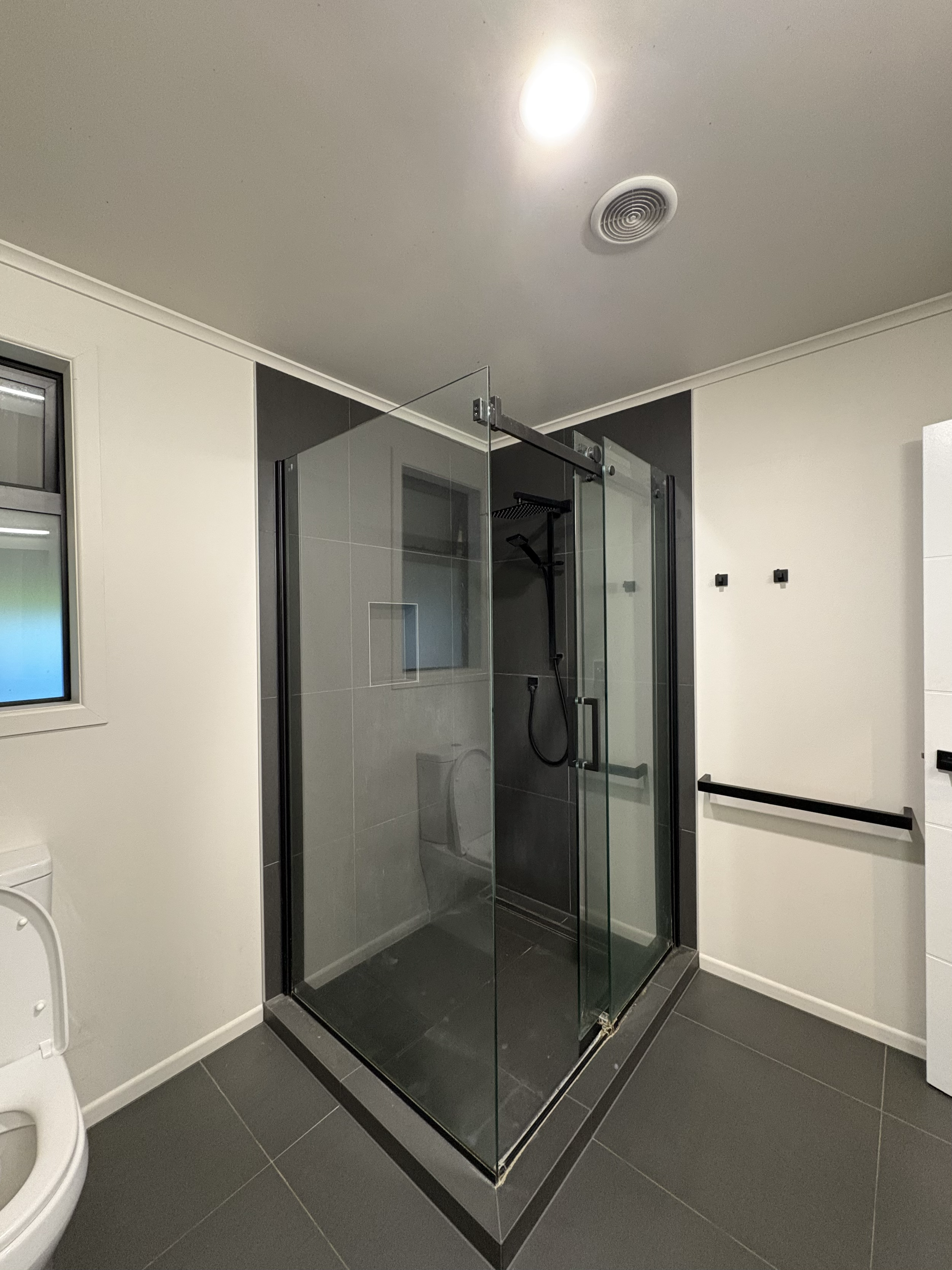 A modern bathroom with a glass shower enclosure, dark tiles, a small window, and a toilet.