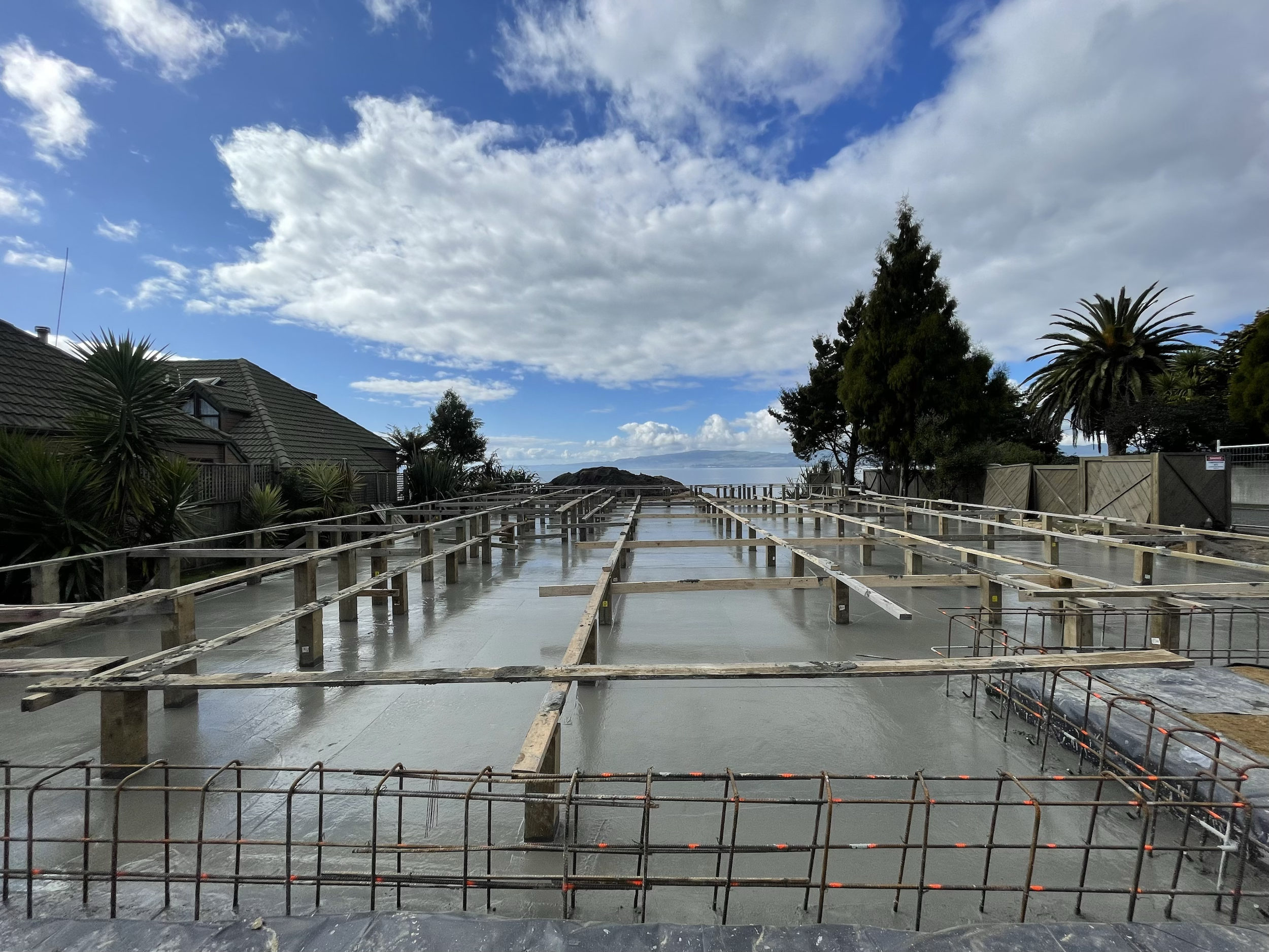 Construction site with concrete foundation and wooden framework in progress, houses on the left, trees on the right, cloudy sky