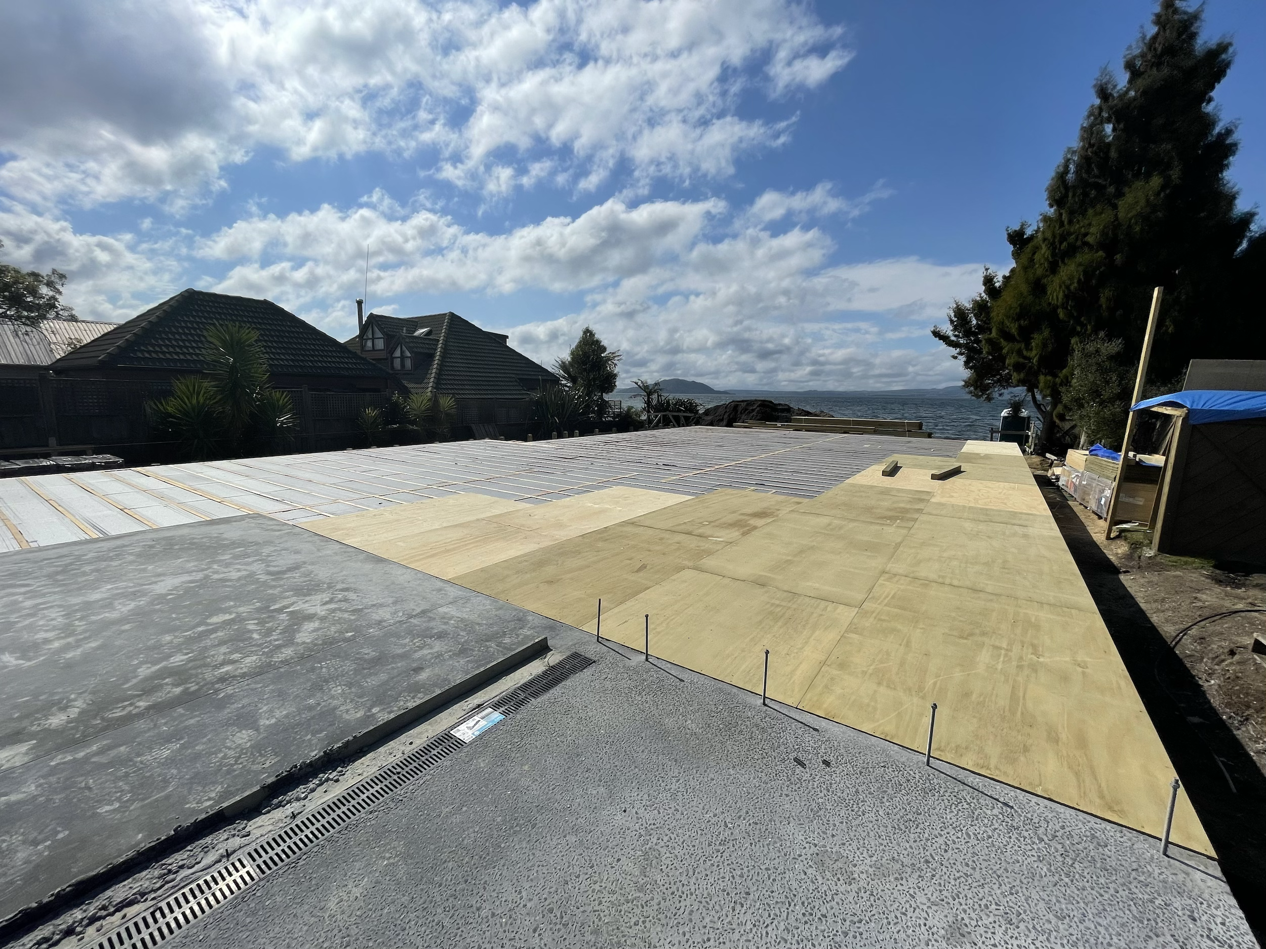 Construction site with plywood and metal sheets on a flat surface, overlooking houses, trees, and the ocean under a partly cloudy sky.