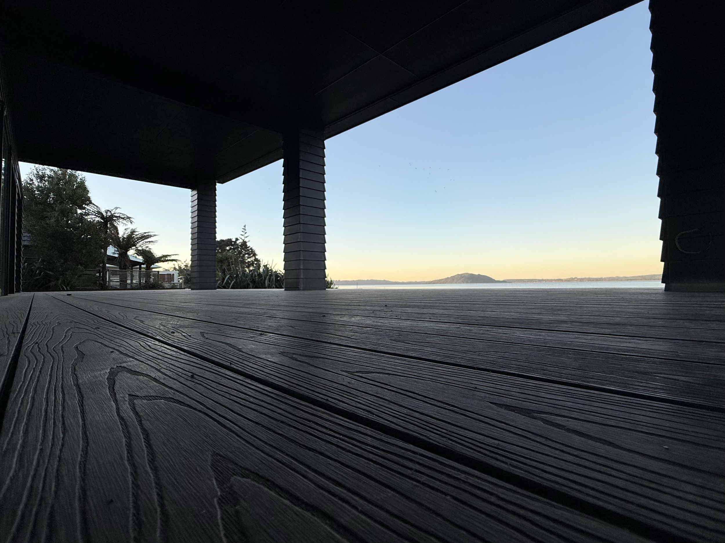 View from a covered wooden deck overlooking a body of water with distant hills, framed by dark support columns and a dark ceiling.