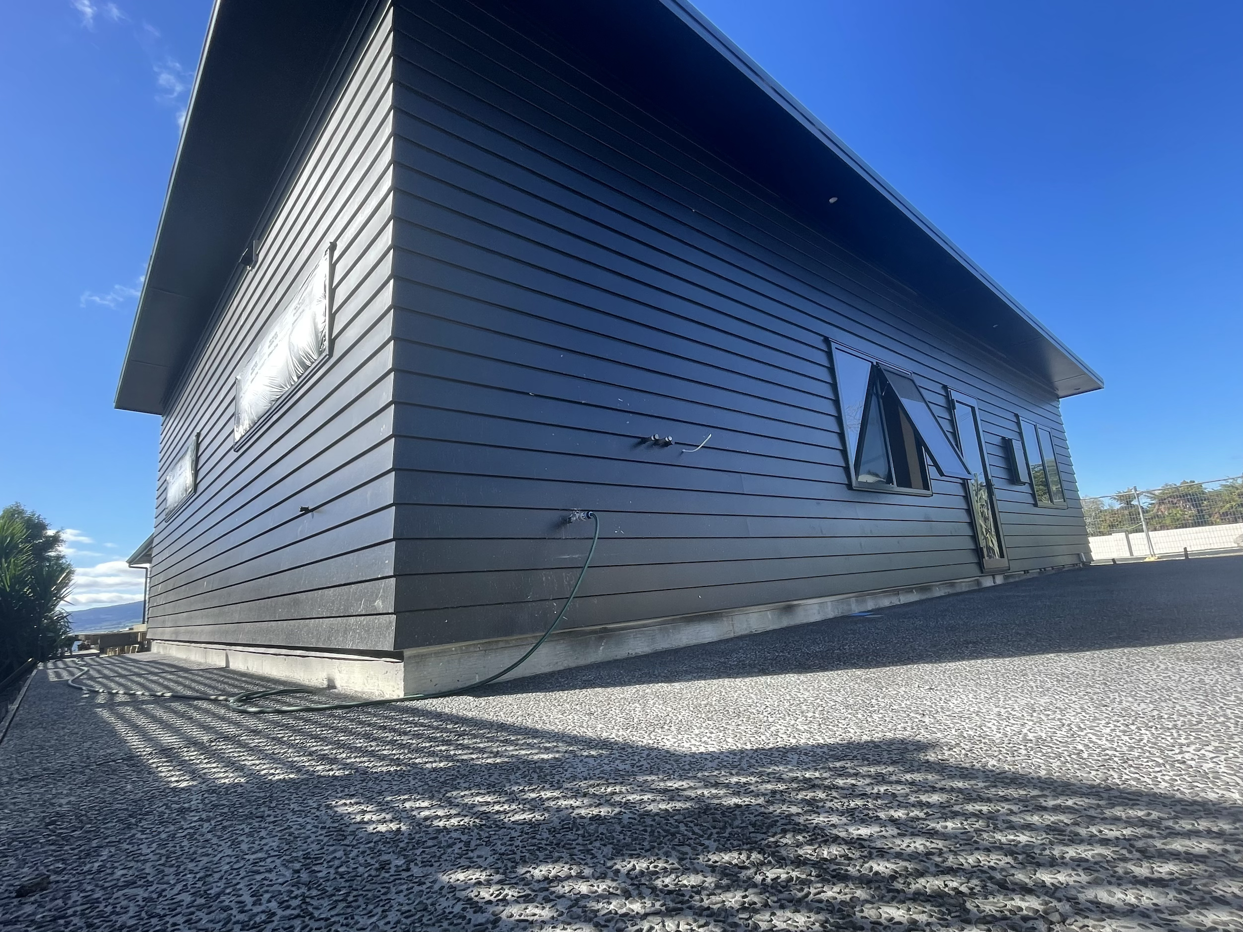 Side view of a modern, black house with angled windows, a flat roof, and a gravel driveway with shadows cast on it.