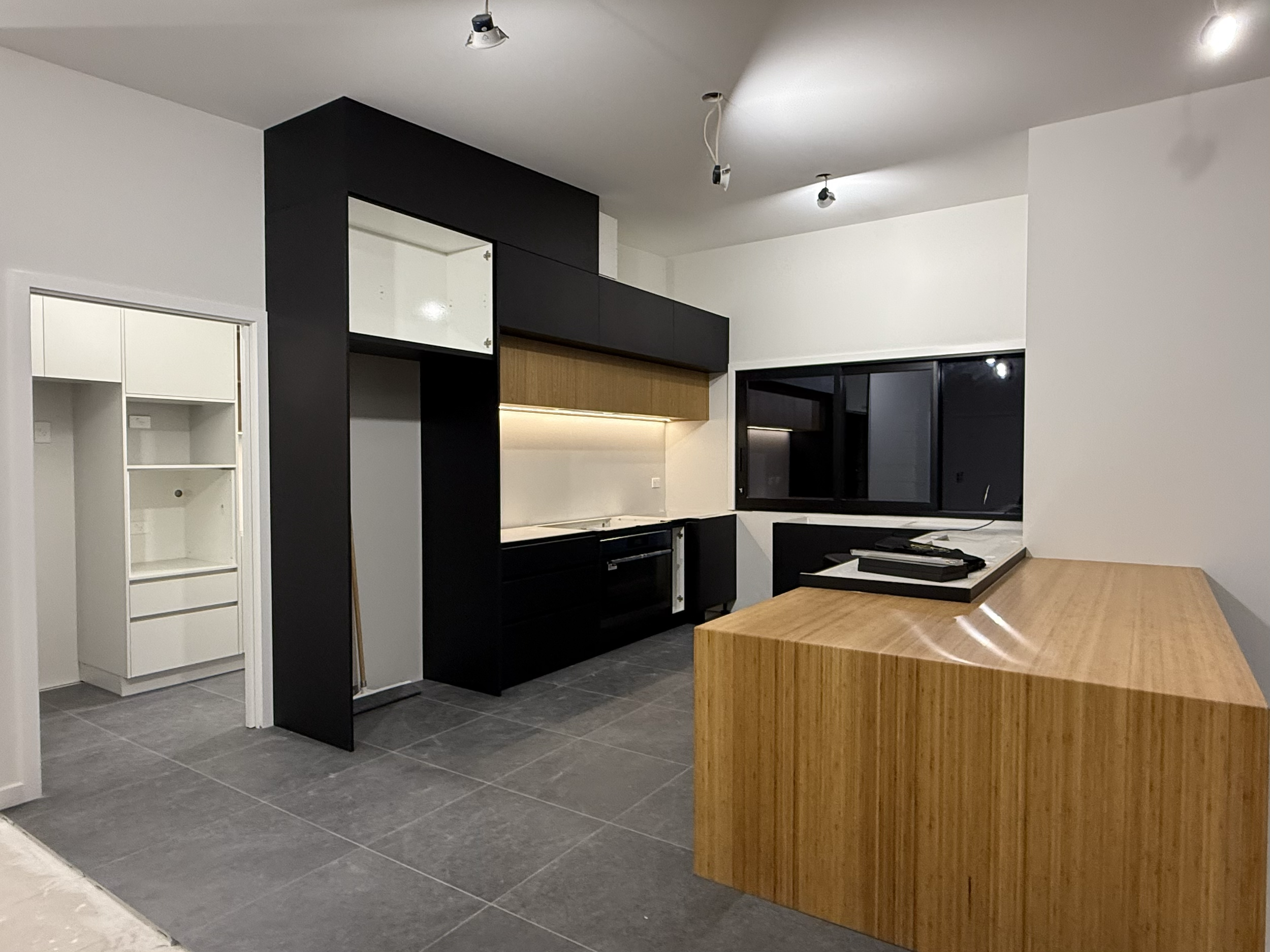 Modern kitchen with black cabinets, a wooden countertop, and gray tiled floor, with a large window and recessed lighting.