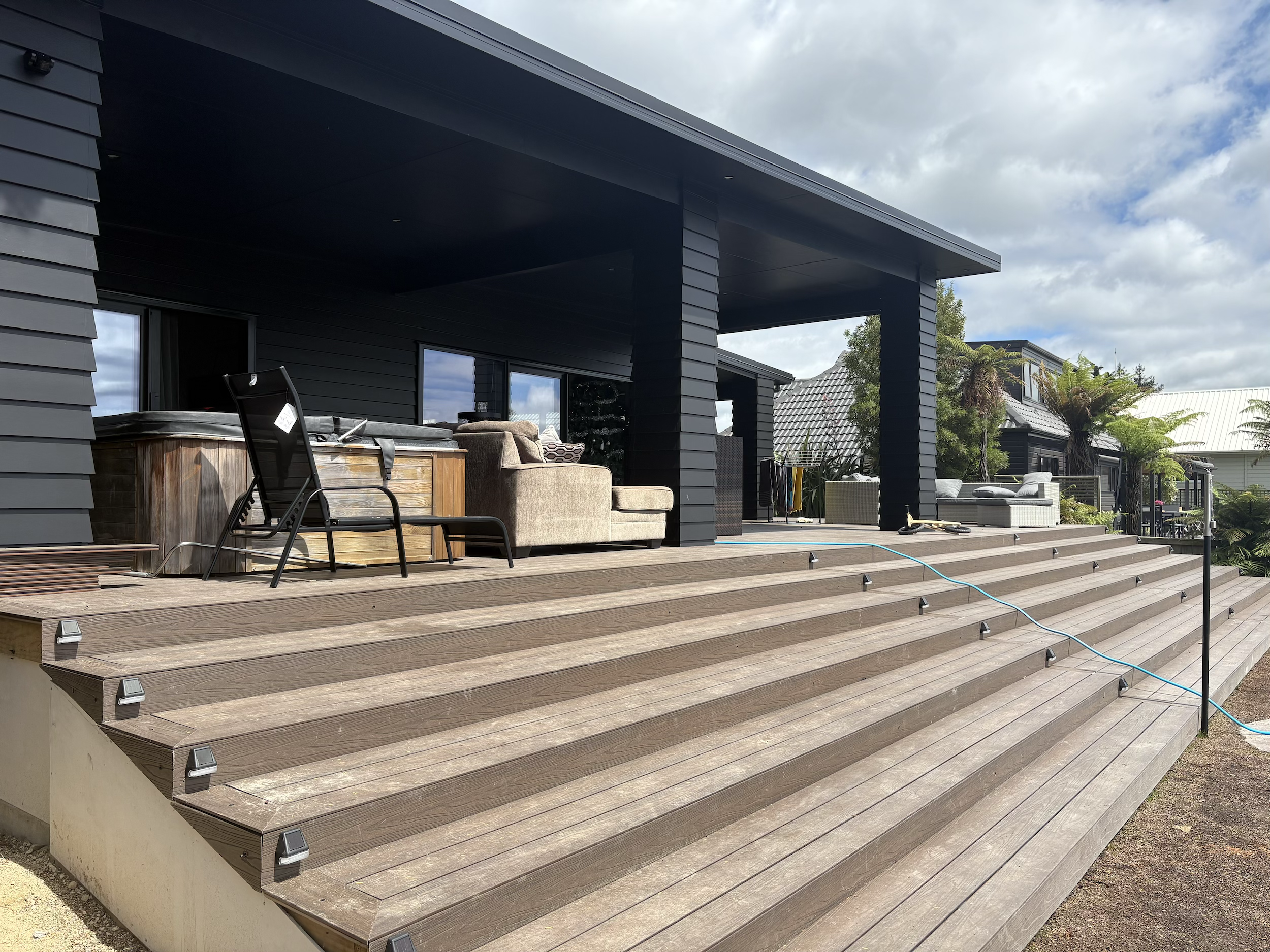 Large wooden outdoor deck with outdoor furniture, including chairs, a hot tub, and sofas, attached to a modern black house with sliding glass doors and a covered patio area, with greenery and other houses in the background under a partly cloudy sky.