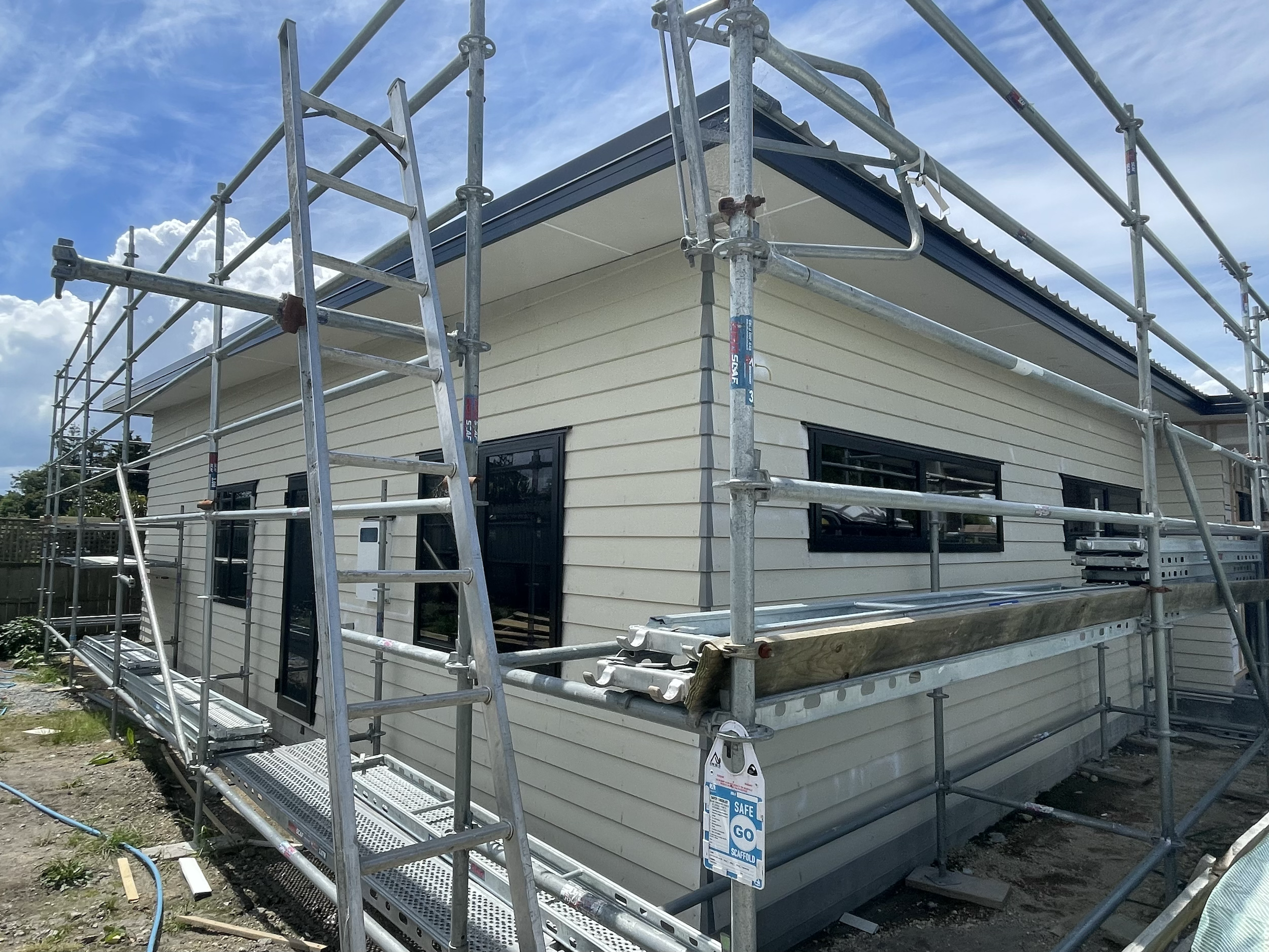 A house under construction with beige siding, black window frames, and scaffolding surrounding it under a blue sky with some clouds.