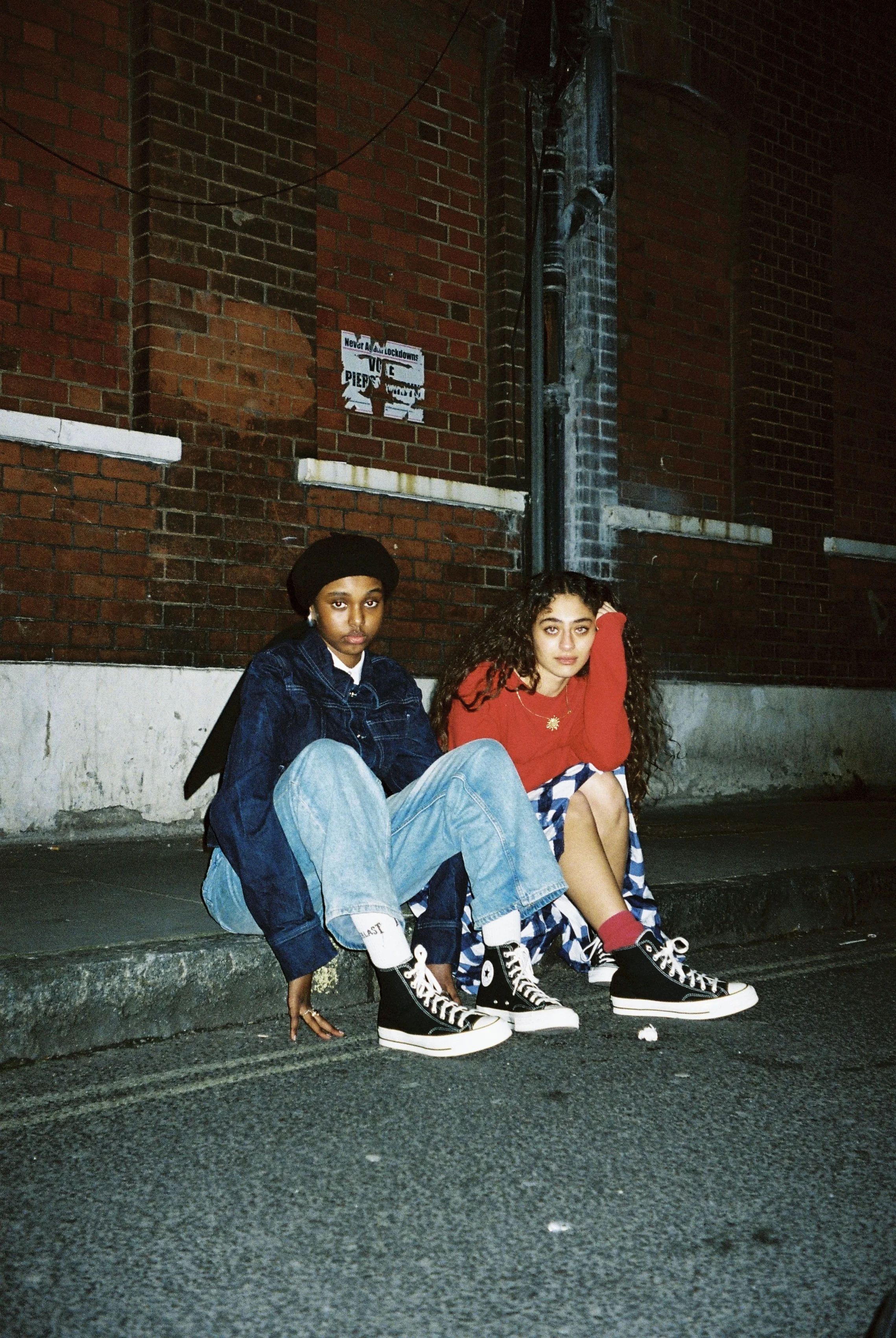 Two young women sitting on a city sidewalk against a brick wall at night. Both are wearing casual clothes and sneakers, with one in a denim jacket and the other in a red sweater and patterned skirt.