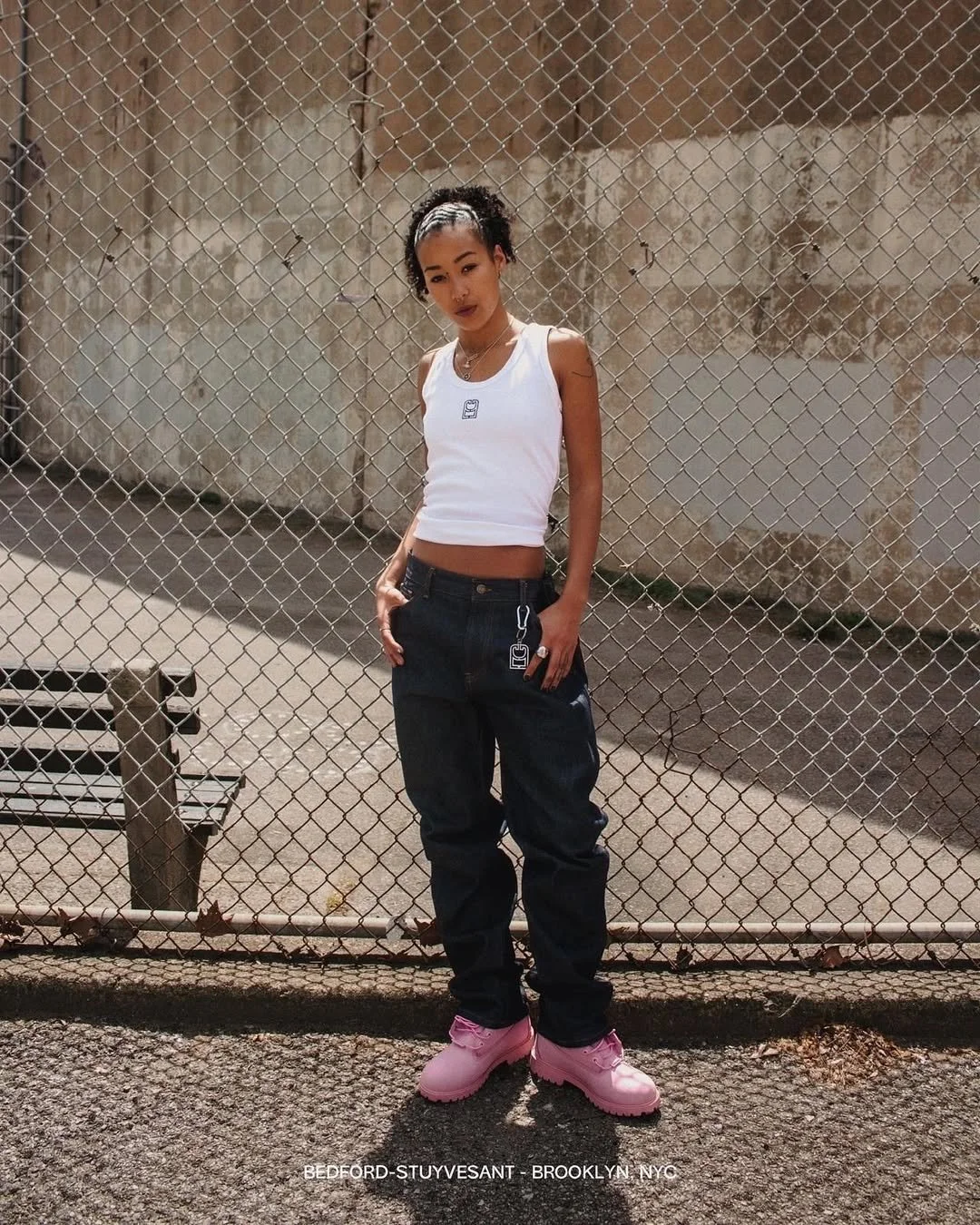 Young woman standing in front of a chain-link fence, wearing a white sleeveless top, dark baggy jeans, and pink boots, with a concrete wall in the background.