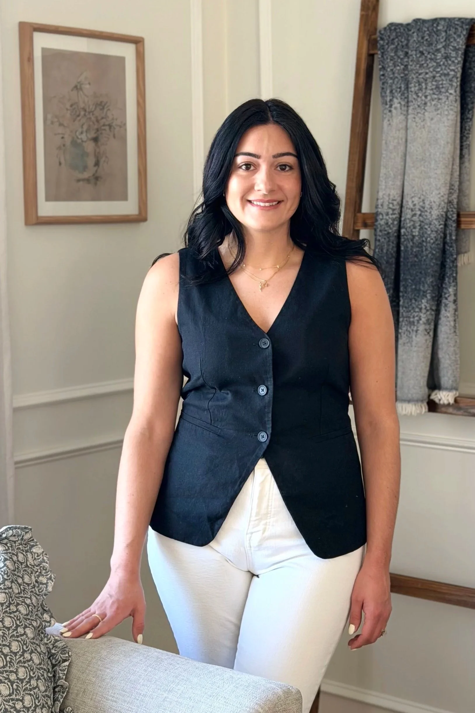 Rochelle of DeMasi Interiors - A woman with black hair wearing a black sleeveless top and white pants, smiling, standing indoors near a beige couch and a decorative pillow.