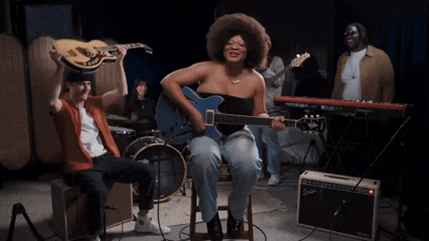 Group of musicians performing in a studio, including a woman singing and playing guitar, a man playing guitar, and a drummer.