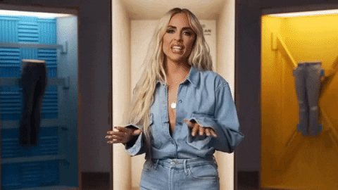 A woman with long blonde hair wearing a light blue shirt and jeans, standing between blue and yellow lockers.