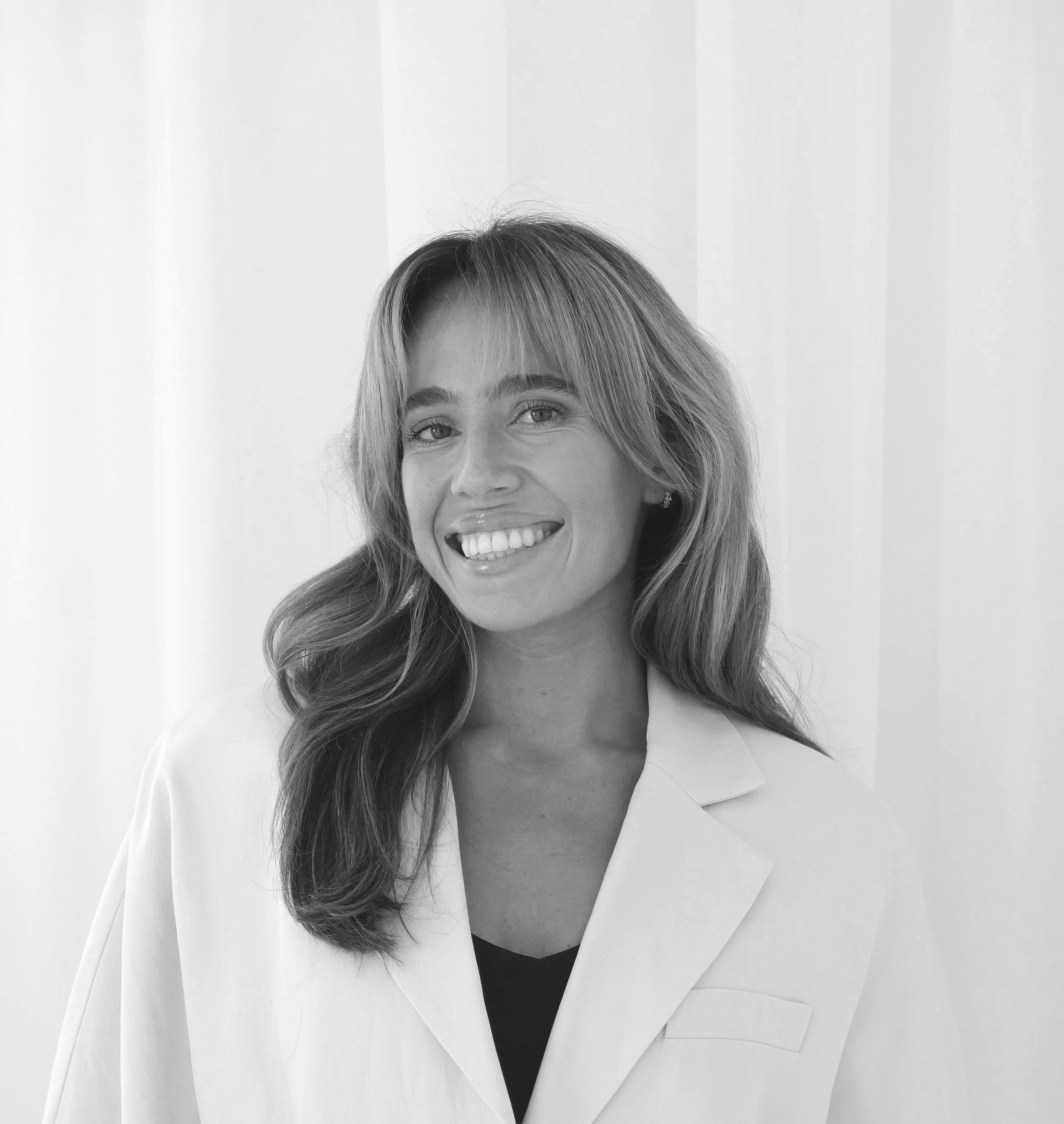 Black and white portrait of a smiling woman with wavy hair wearing a light-colored blazer over a dark top, standing in front of a plain background.
