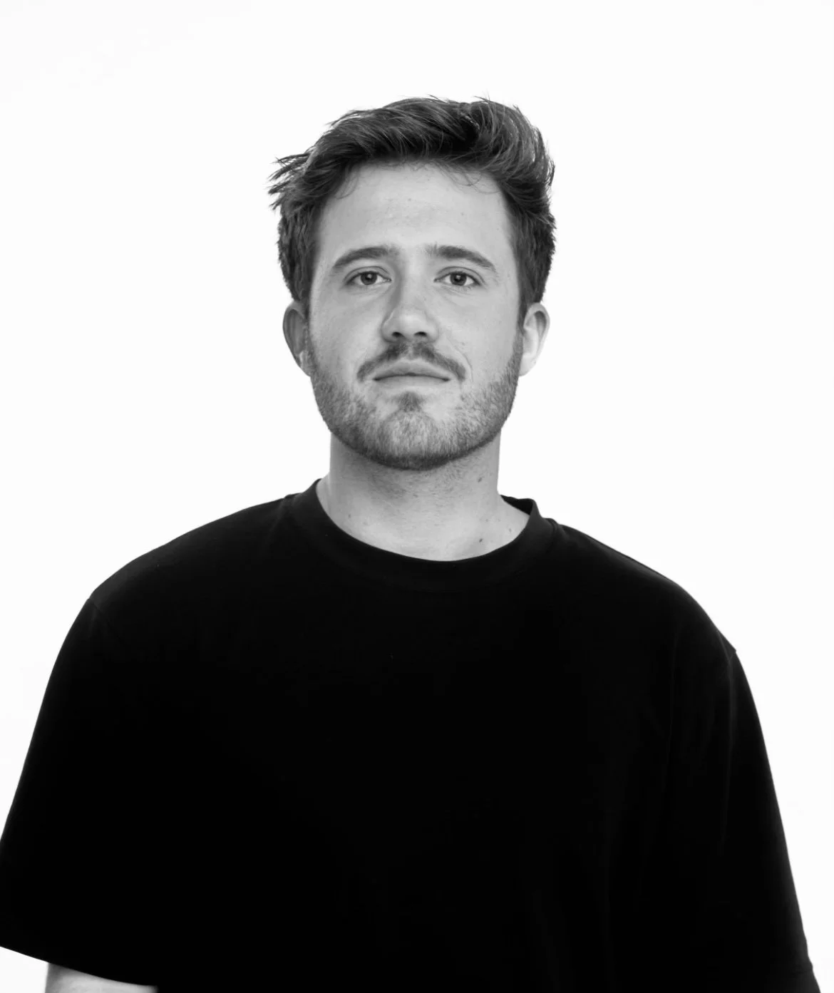 Black and white photo of a young man with a beard and styled hair, wearing a black T-shirt, looking directly at the camera against a blank background.