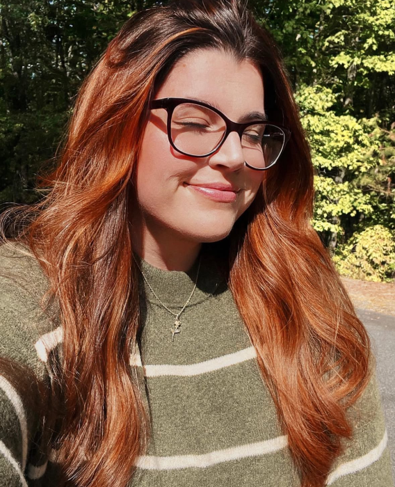Young woman with long, wavy auburn hair smiling with eyes closed, wearing glasses and a green striped sweater outdoors with trees in the background. the social porch hayesville north carolina