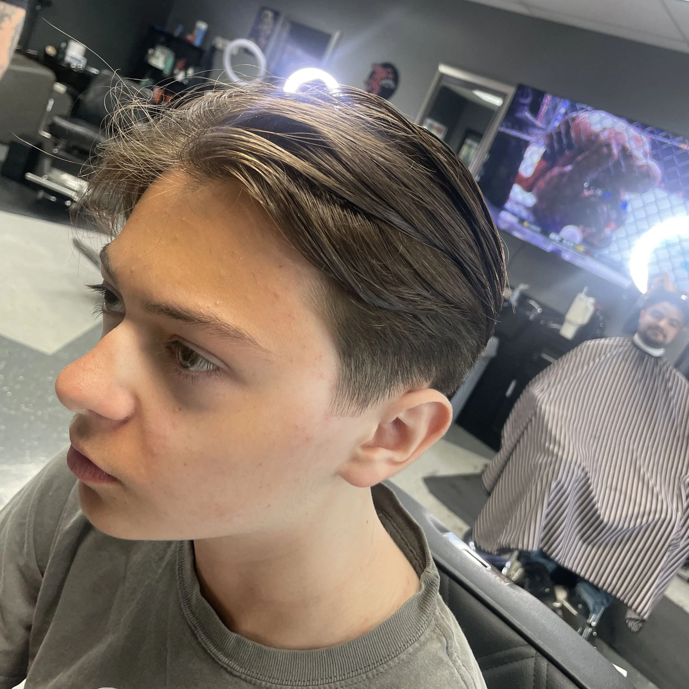 Young man with a fresh, styled haircut sitting in a barbershop, with barber in the background and a television screen displaying a fight scene.