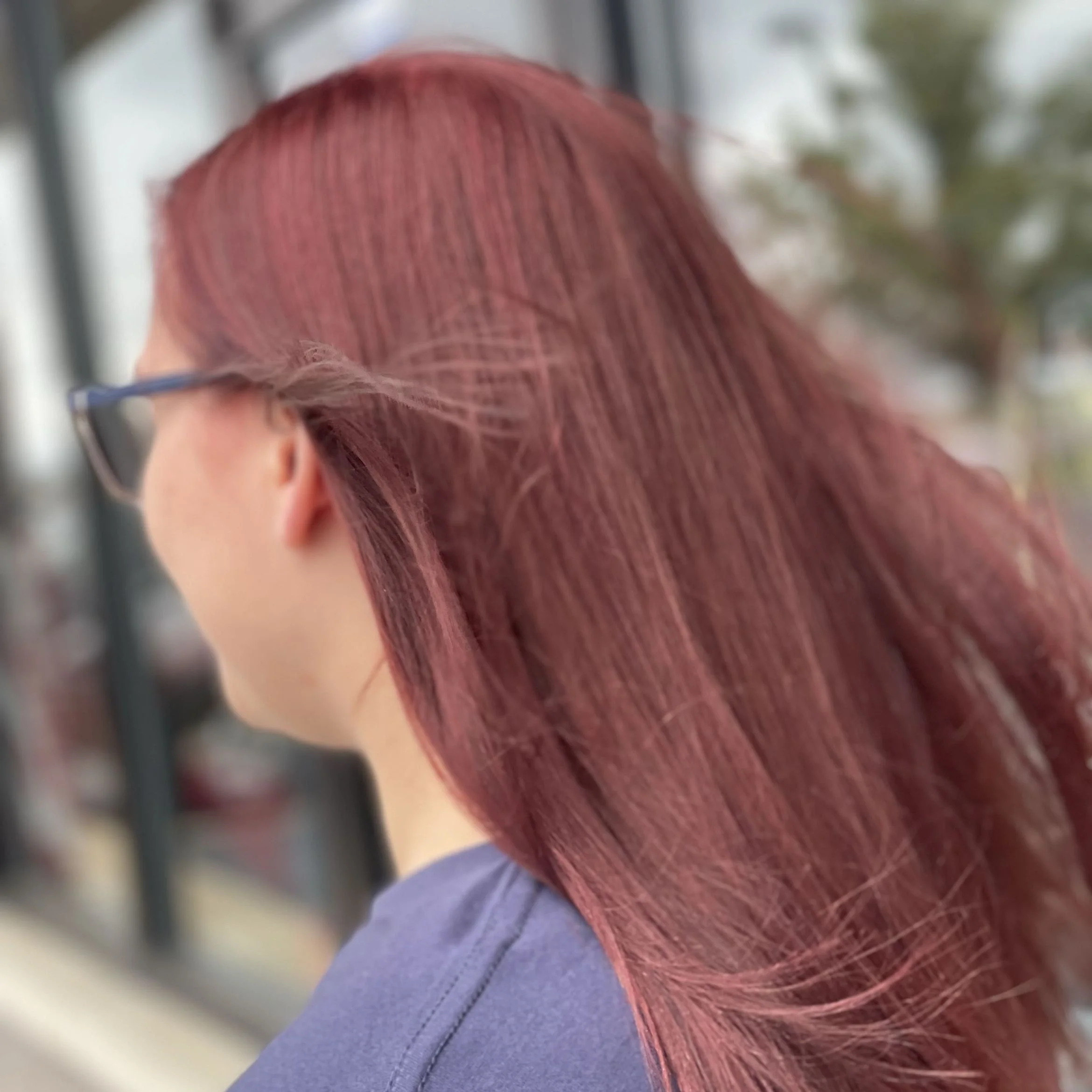 Rear side view of a woman with reddish-brown hair and glasses, outdoors near a glass window.