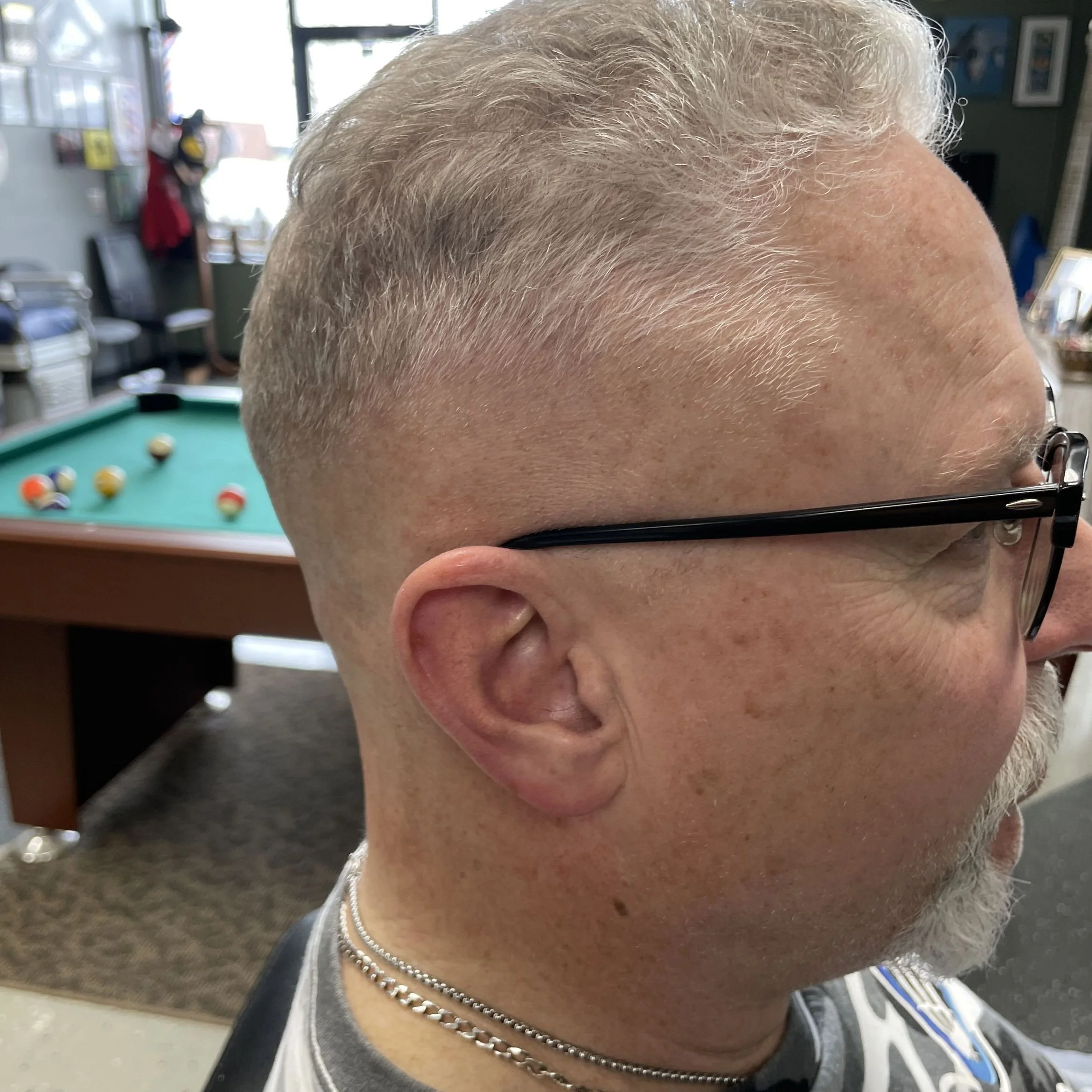 Close-up of a man with light gray hair, wearing glasses and silver necklaces, in an indoor setting with a pool table in the background.