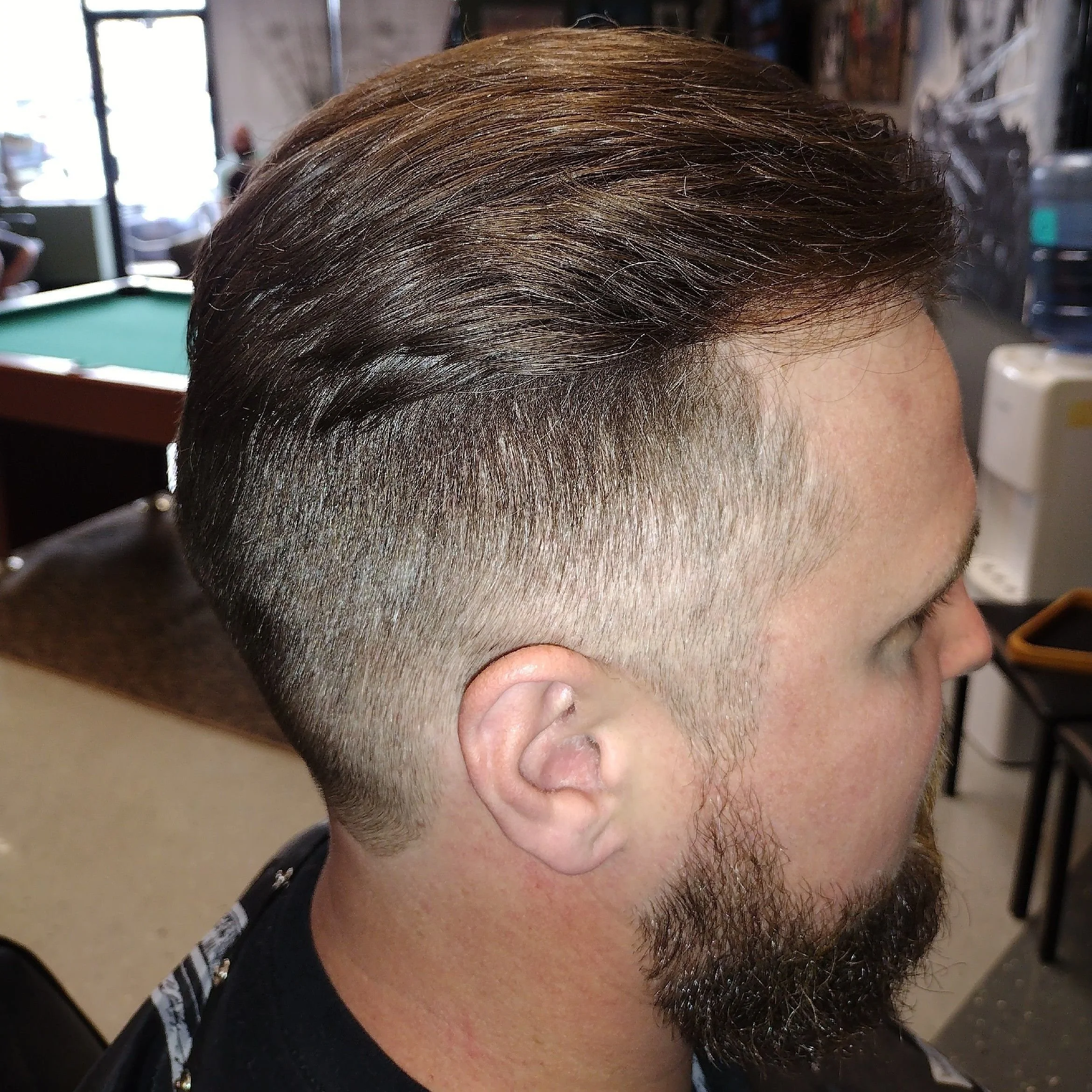 Side view of a man with a modern hairstyle, featuring a fade on the sides and longer, slicked-back hair on top, in a barbershop setting.