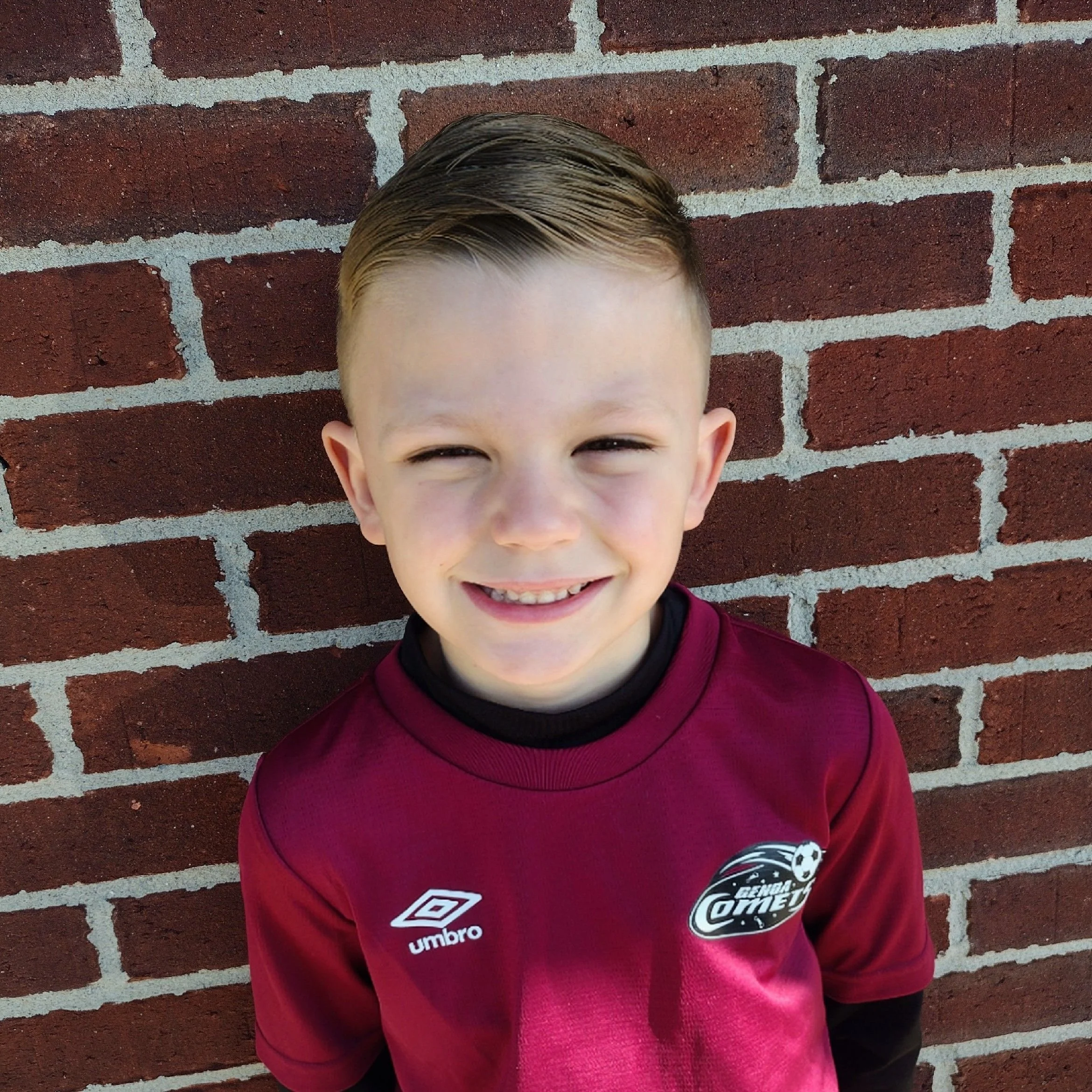 Smiling young boy with short brown hair standing against a red brick wall, wearing a maroon soccer jersey with the Umbro logo and a team badge.