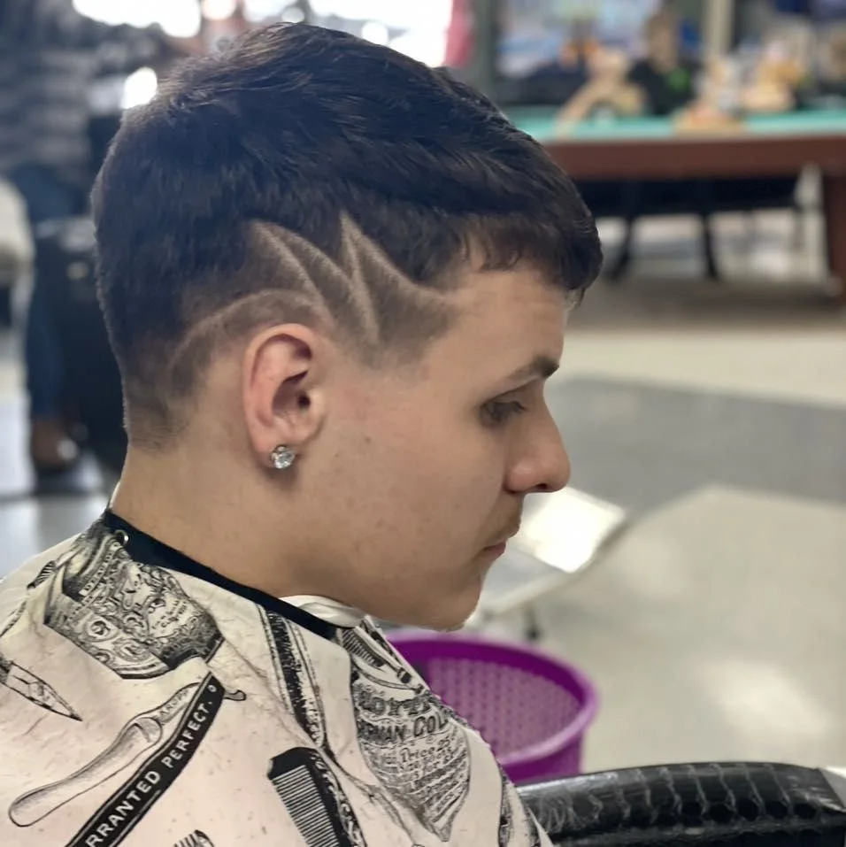 A young man with short, dark hair and a shaved design on the side, wearing a patterned cape and earrings, sitting in what appears to be a barbershop.