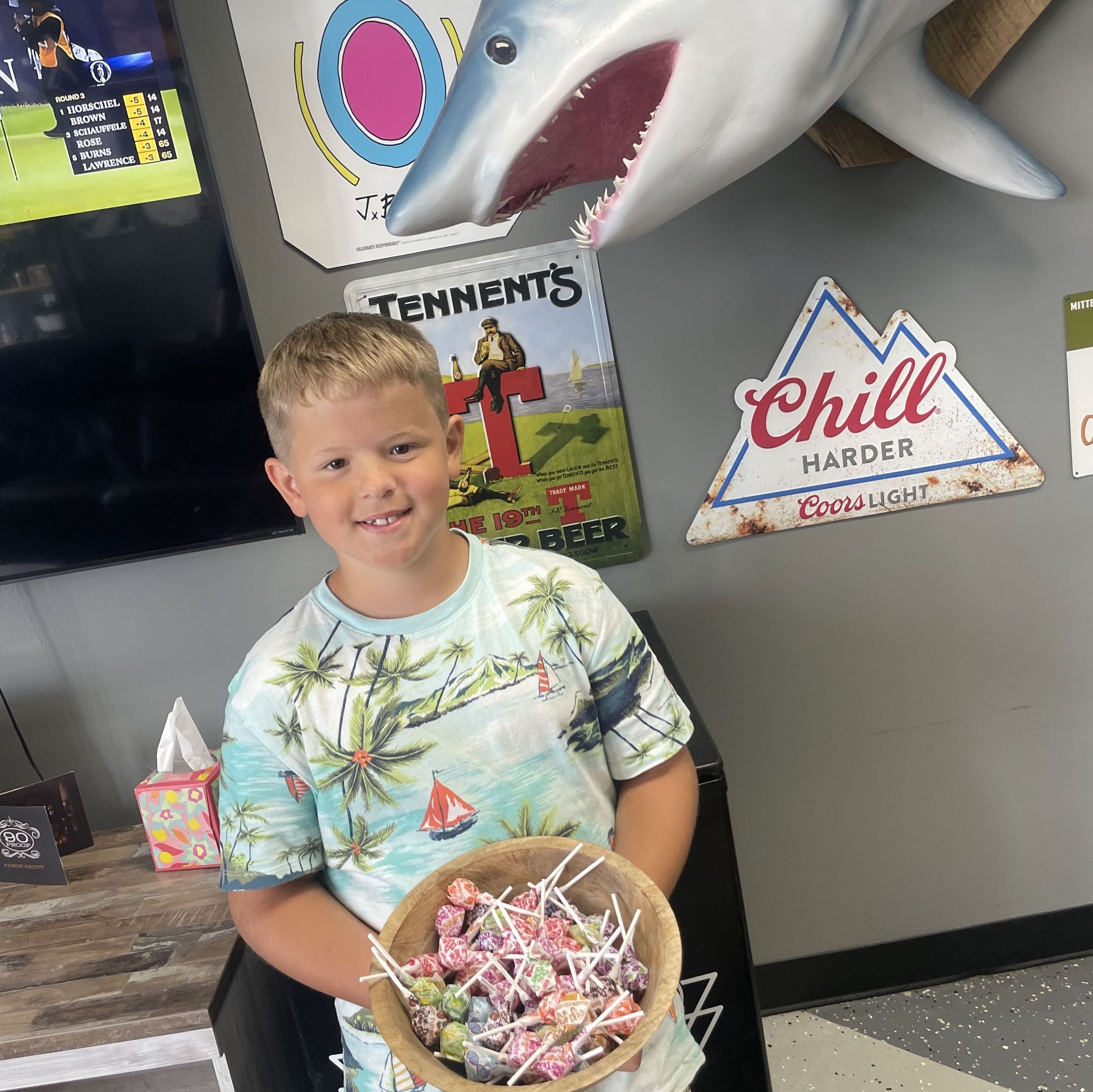 A young boy holding a bowl of colorful candy with lollipops at a casual indoor setting. The background features various posters and signs, including a shark head sculpture, a sports TV screen, and a Tennent's beer sign.