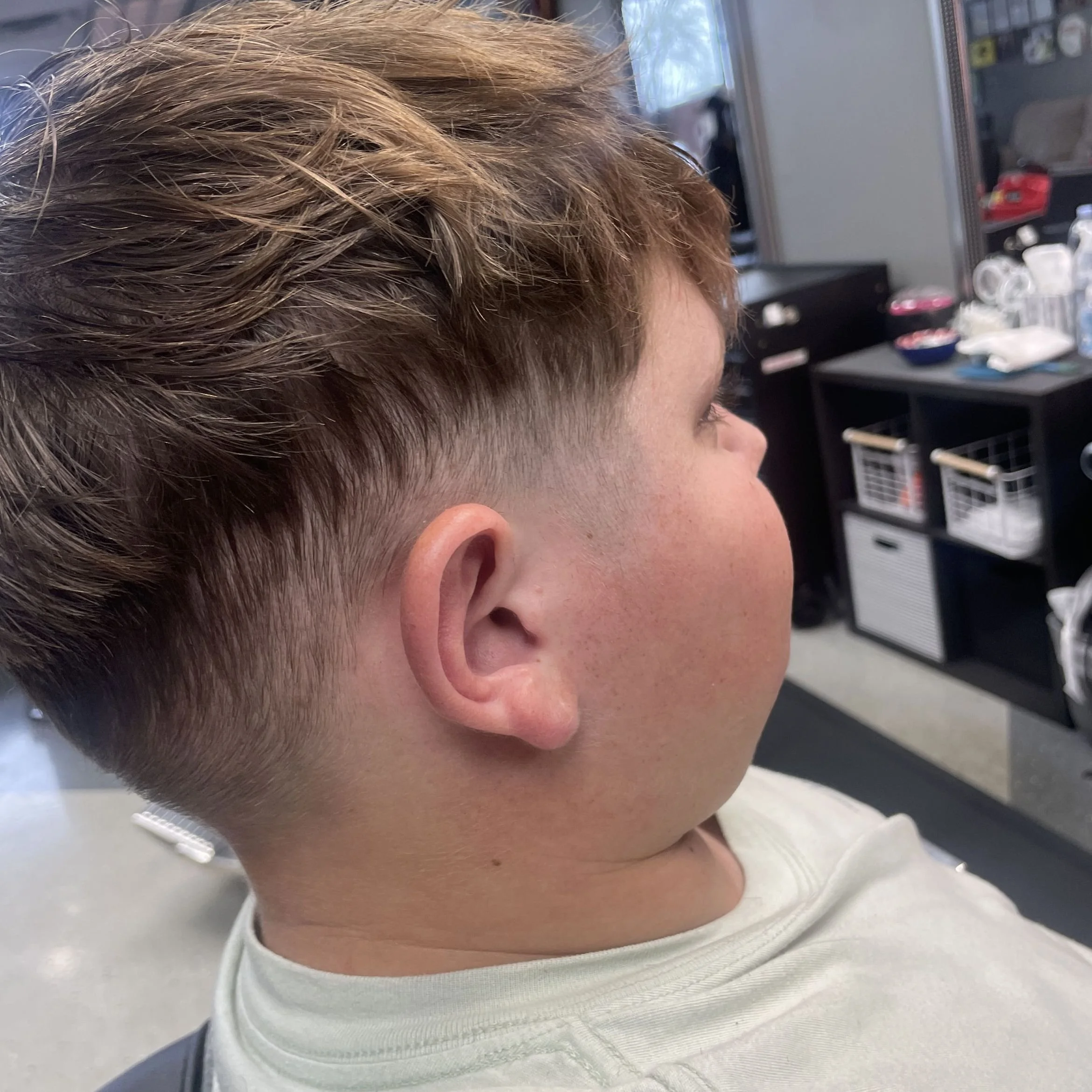 Close-up side view of a young person with a fresh haircut, short on the sides and longer on top, sitting in a salon or barbershop with hairstyling tools and supplies in the background.