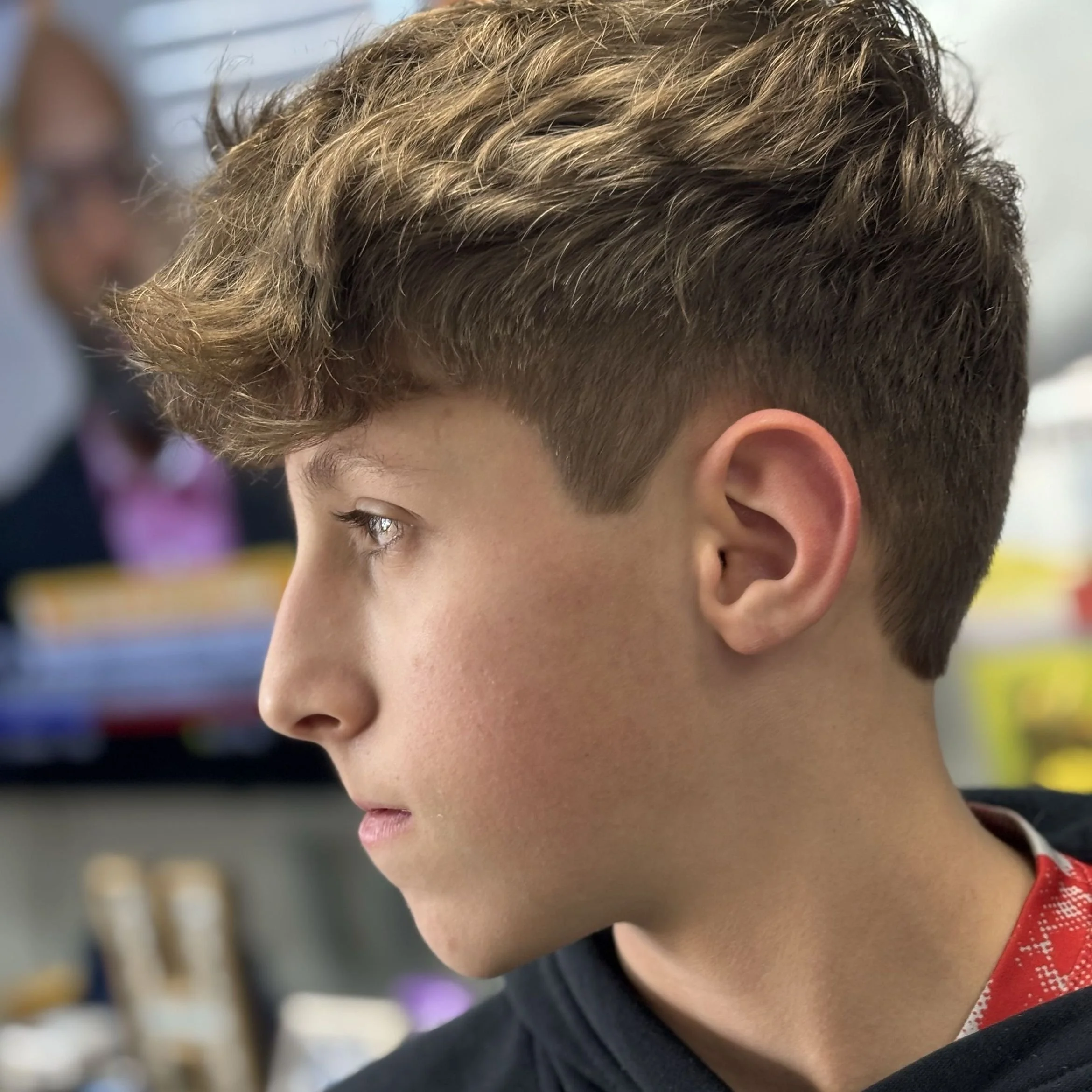 Close-up side profile of a boy with brown curly hair, wearing a black shirt, indoors with other people in the background.